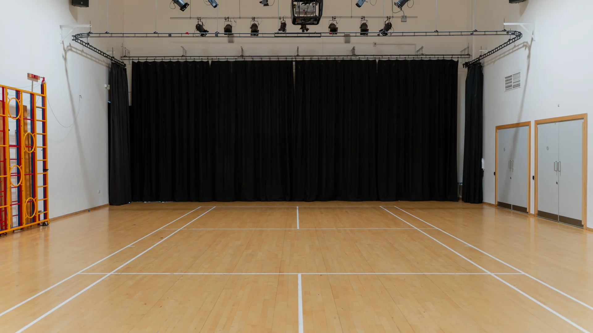 A photo of the Main Hall at Deptford Lounge. It looks like a school sports hall, with court lines on the floor, and gymnastics equipment along one wall. There is a lighting rig on the ceiling and a black curtain covering the back wall.