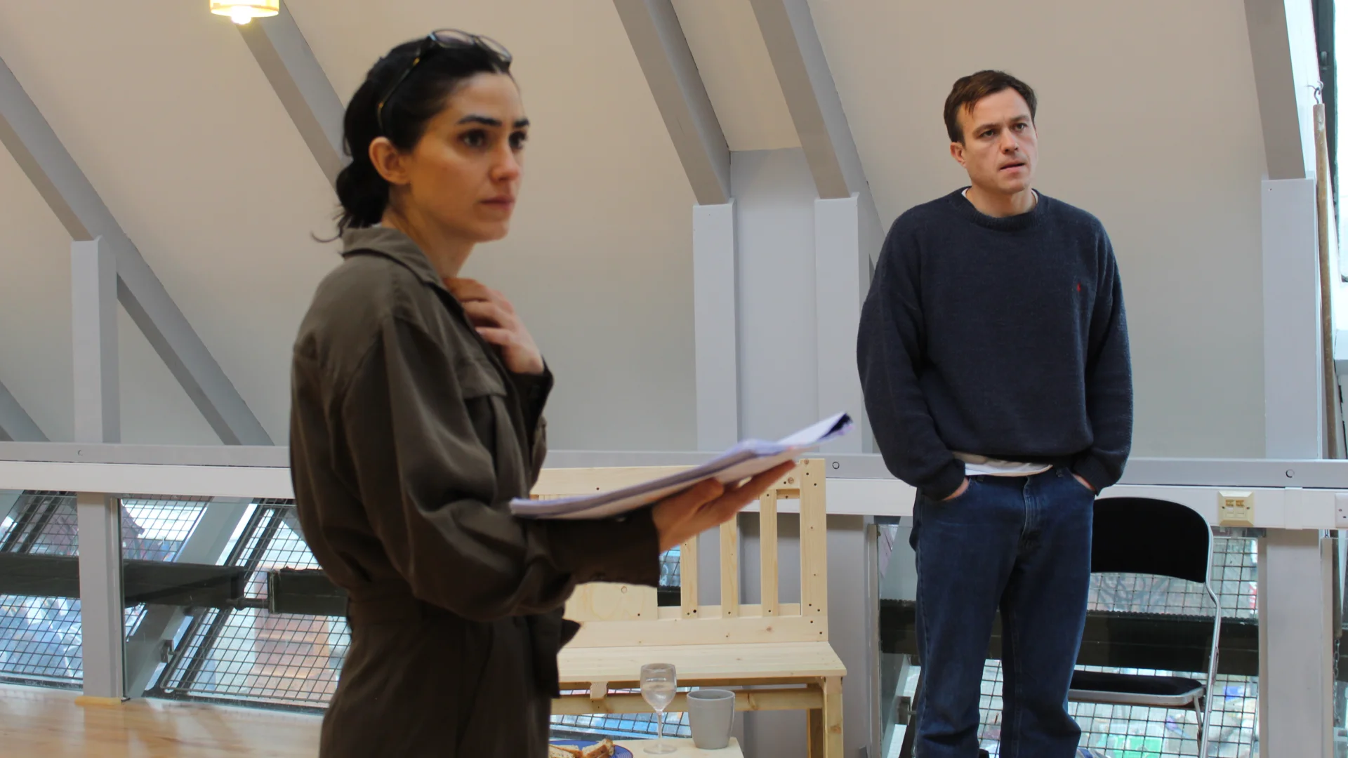 Cassie Bradley and Barney White in rehearsal for One Day When We Were Young. A woman wearing a grey boiler suit stands with one hand holding a script and the other on her chest. Behind her is a man wearing jeans and a black jumper with his hands in his pockets.