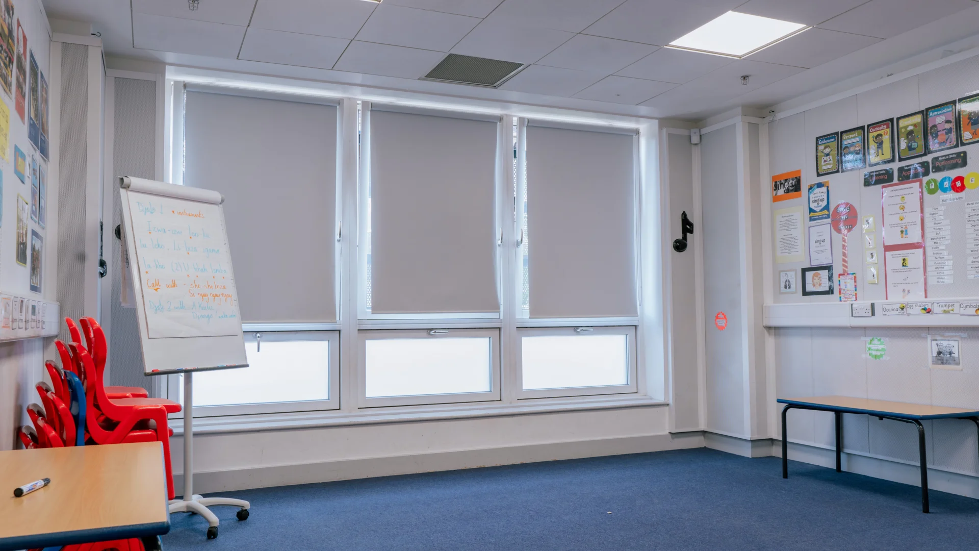 A photo of the music room at Deptford Lounge. It is a room with white walls and a blue carpeted floor. One wall has floor to ceiling windows with the blinds pulled most of the way down. In the space is a flipchart, two tables and a stack of orange and black chairs.