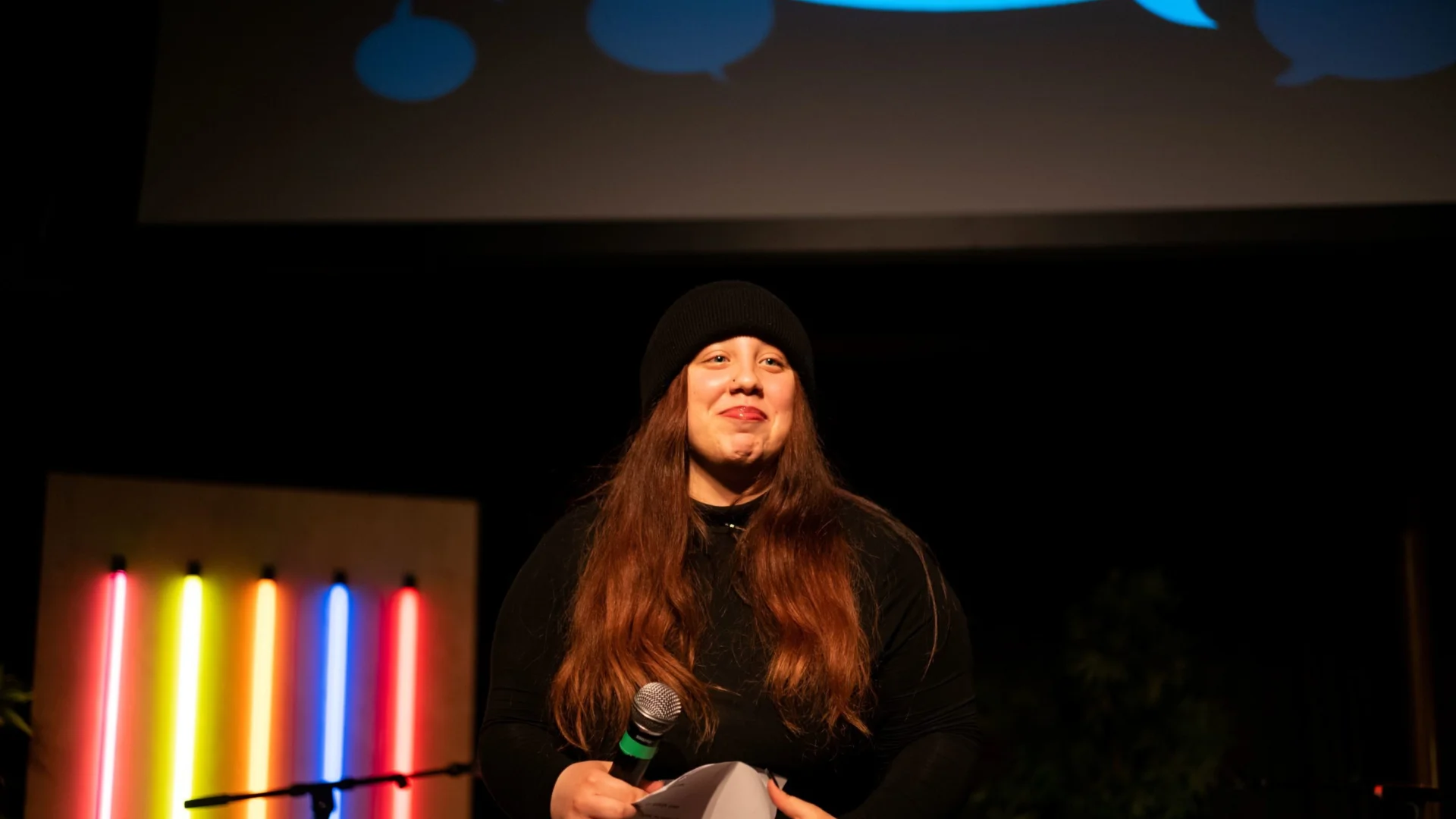 A person in a black outfit and long brown hair wearing a beanie holds a microphone on stage with a colourful neon light display behind them, conveying a bright and lively atmosphere.