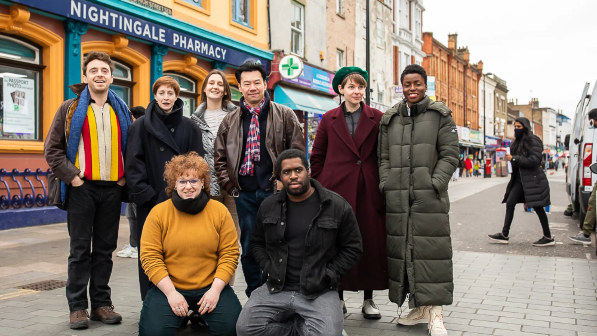 A diverse group of eight people, dressed warmly, gather on a lively Deptford High Street." The mood is cheerful.