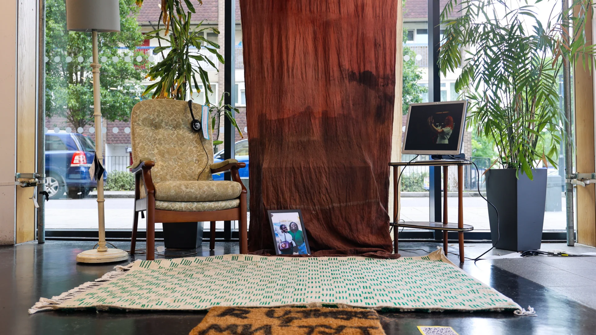 A photo of an installation of a living room. On the floor there is a rug, an armchair, a lamp, a TV, pot plants and a framed photo.