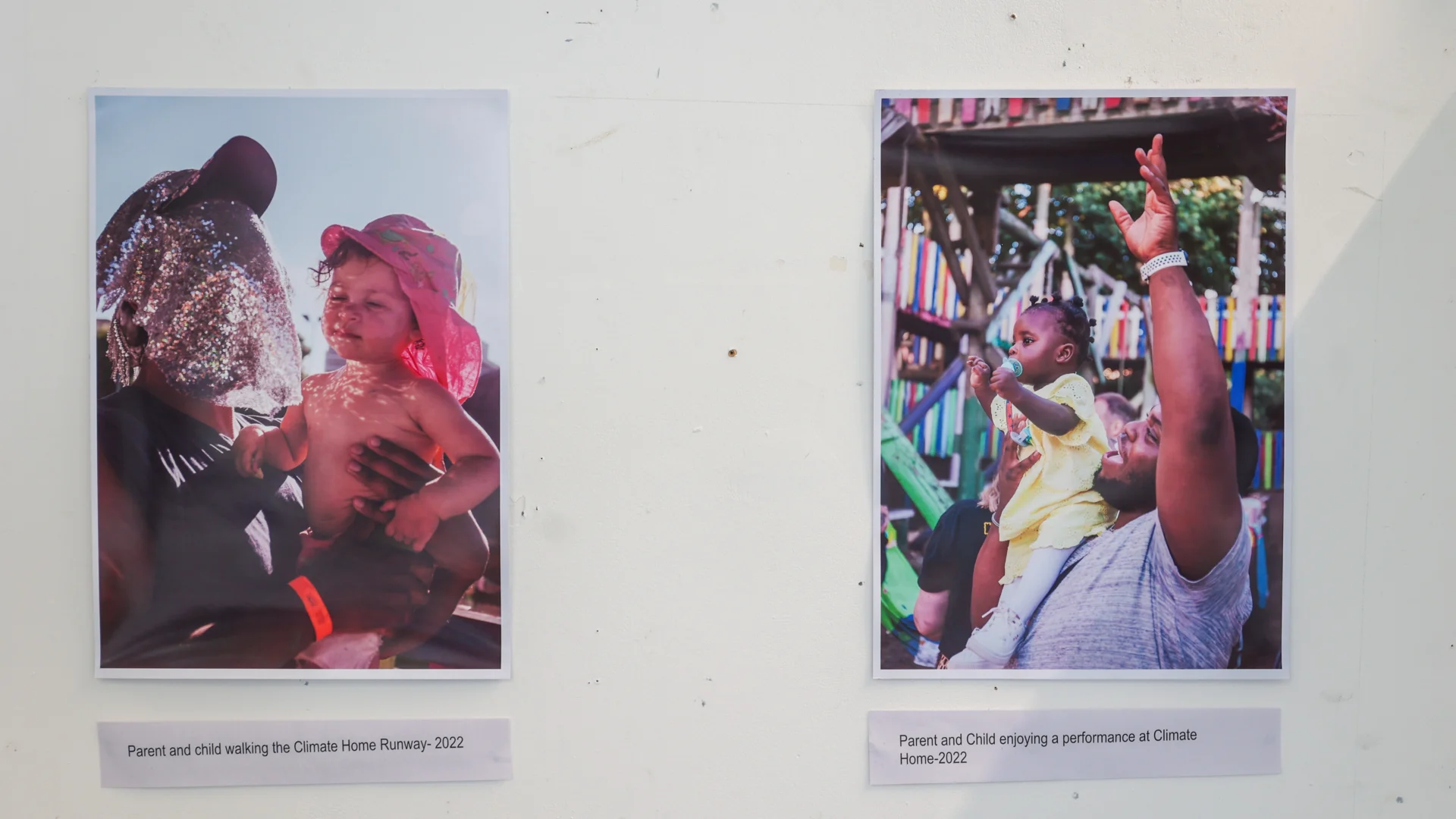 A photograph of a white exhibition wall with two printed photos mounted on it. The first is of a black person wearing a glittery piece of fabric over their face, holding a baby wearing a pink hat. The second is of a black man holding a baby wearing a yellow outfit. The man has his arm raised to the sky.