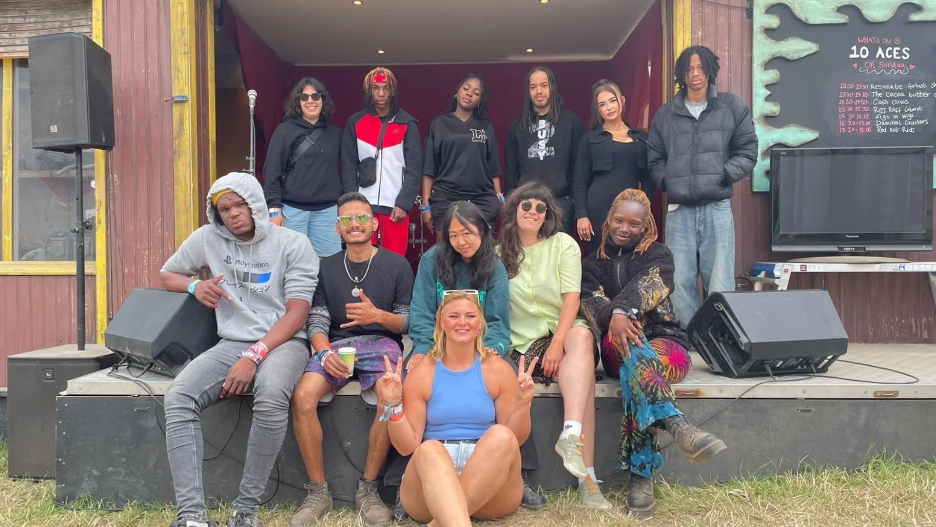 A diverse group of young people pose happily on a wooden stage outdoors. Some sit on the edge while others stand in a line behind them. The mood is relaxed and joyful.
