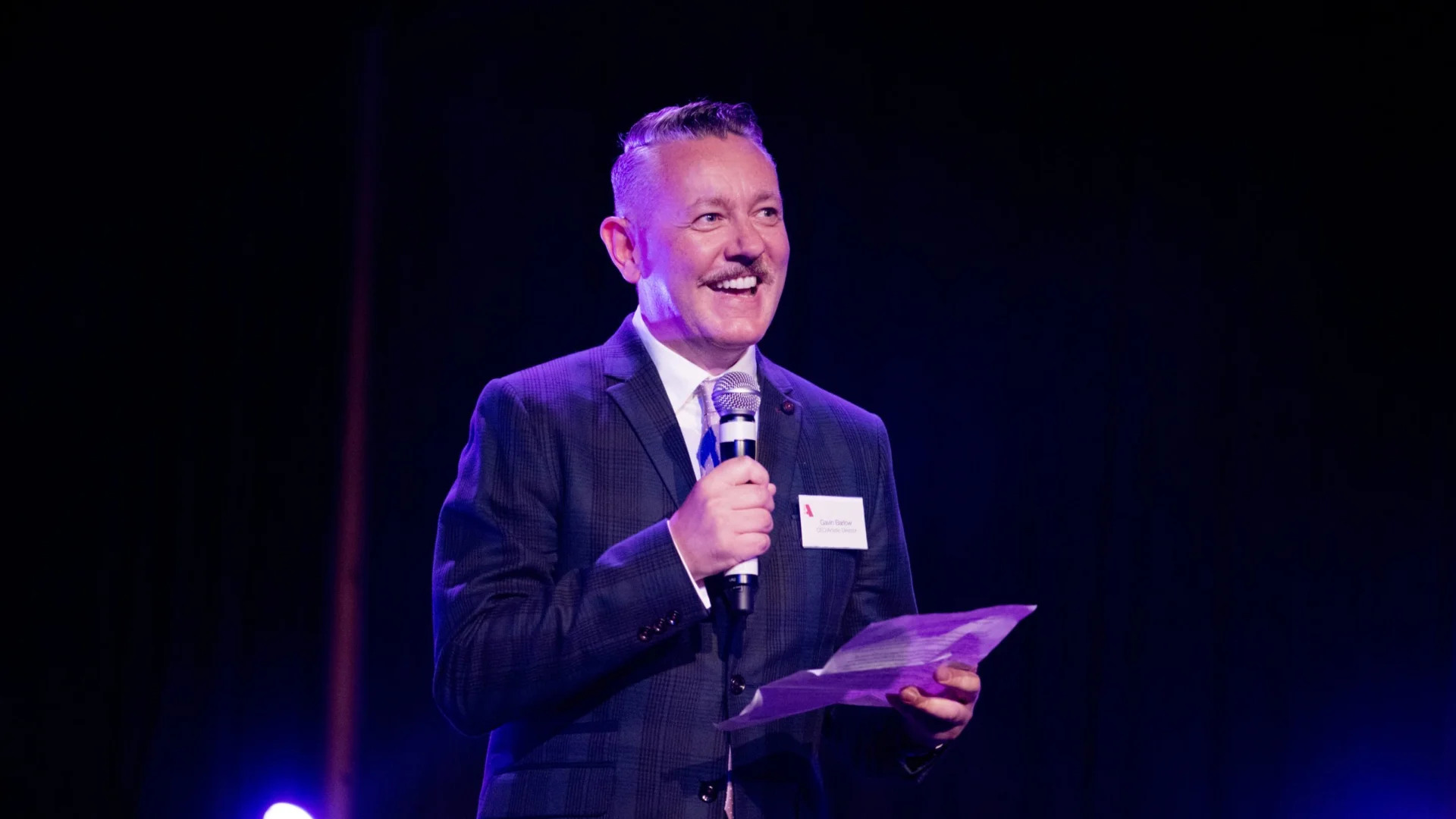 A man in a suit is smiling and holding a microphone on a dark stage. He is uplift in pink and blue, the mood is joyful and celebratory.