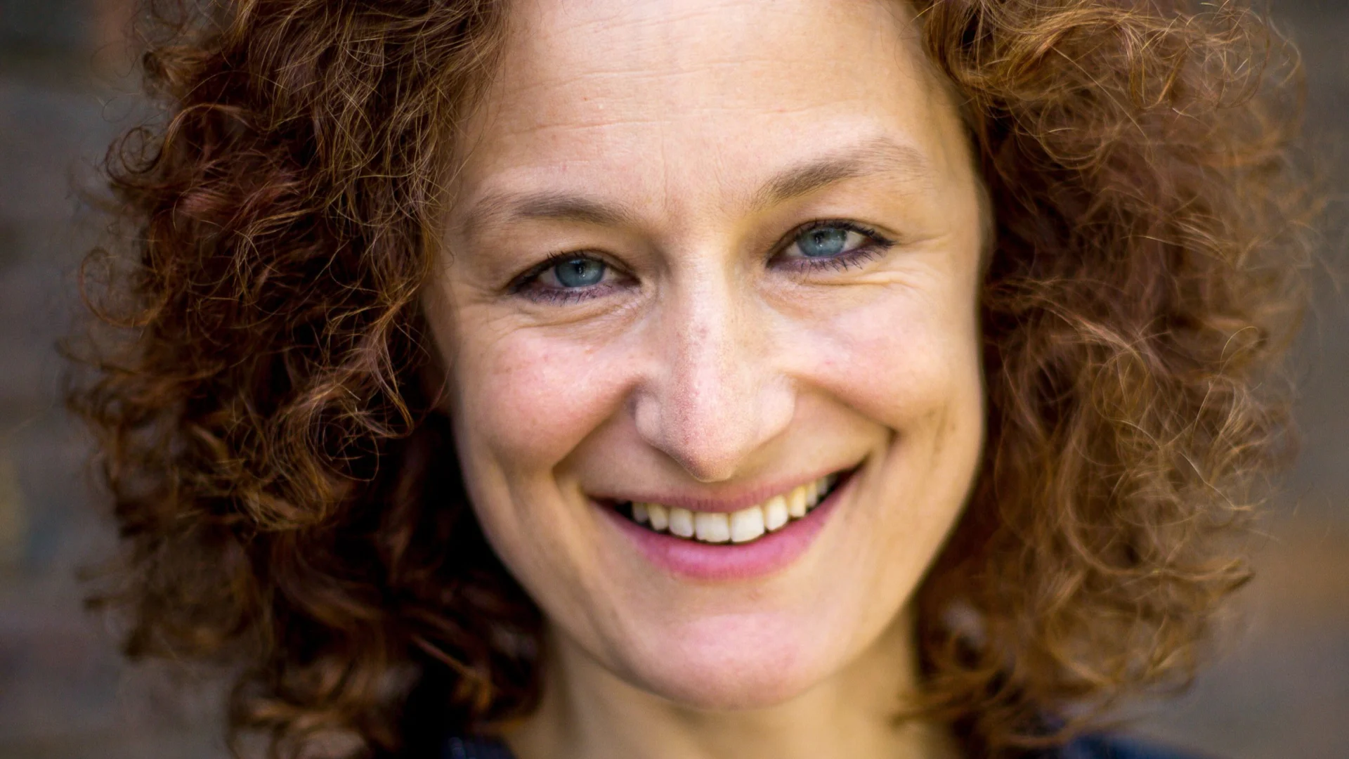 A woman with curly red hair and a friendly smile is wearing a black shirt. She stands against a blurred brick wall, conveying warmth and approachability.