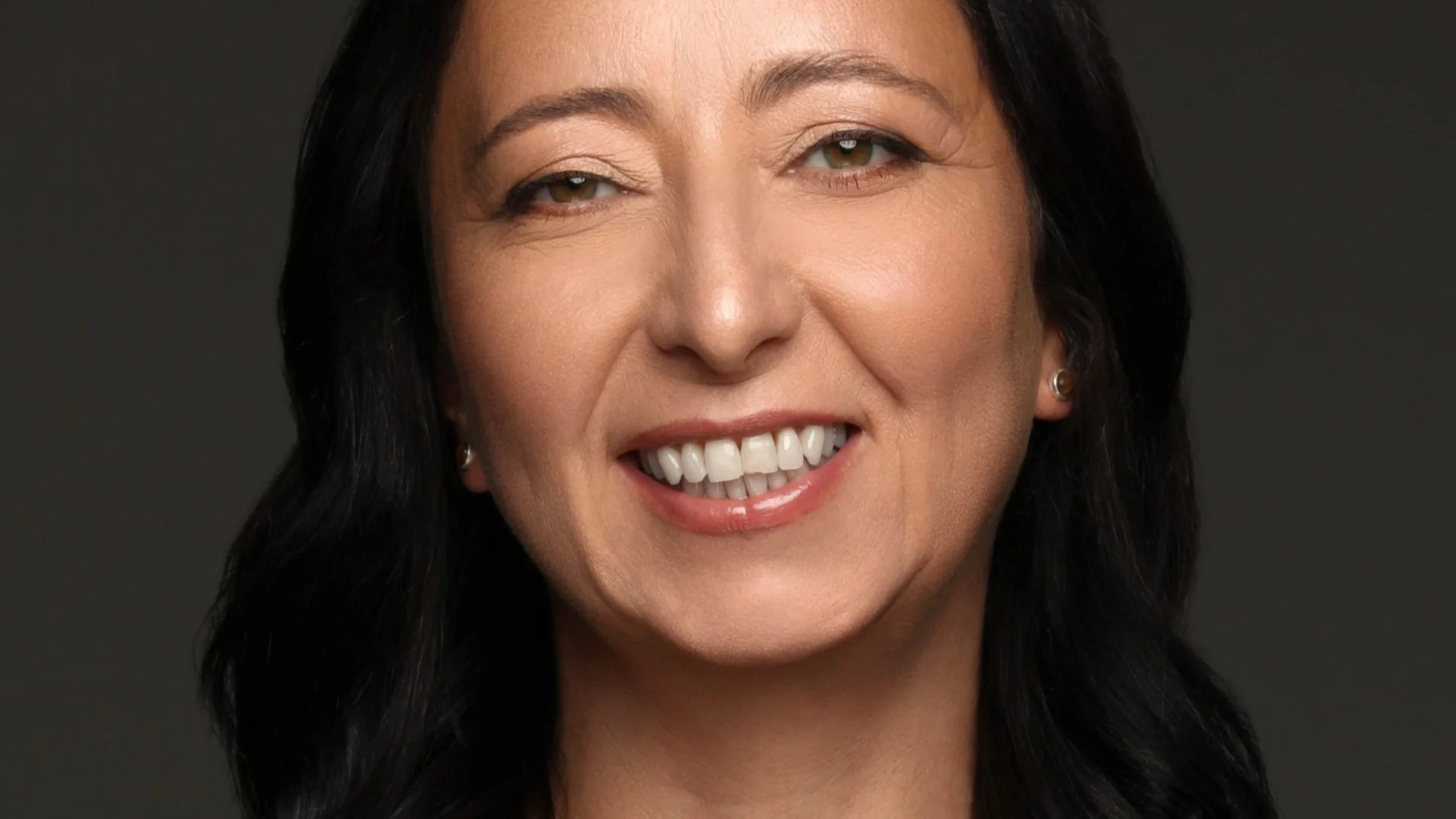Smiling woman with shoulder-length black hair, wearing a teal top and a simple necklace, against a plain dark background. She conveys warmth and confidence.