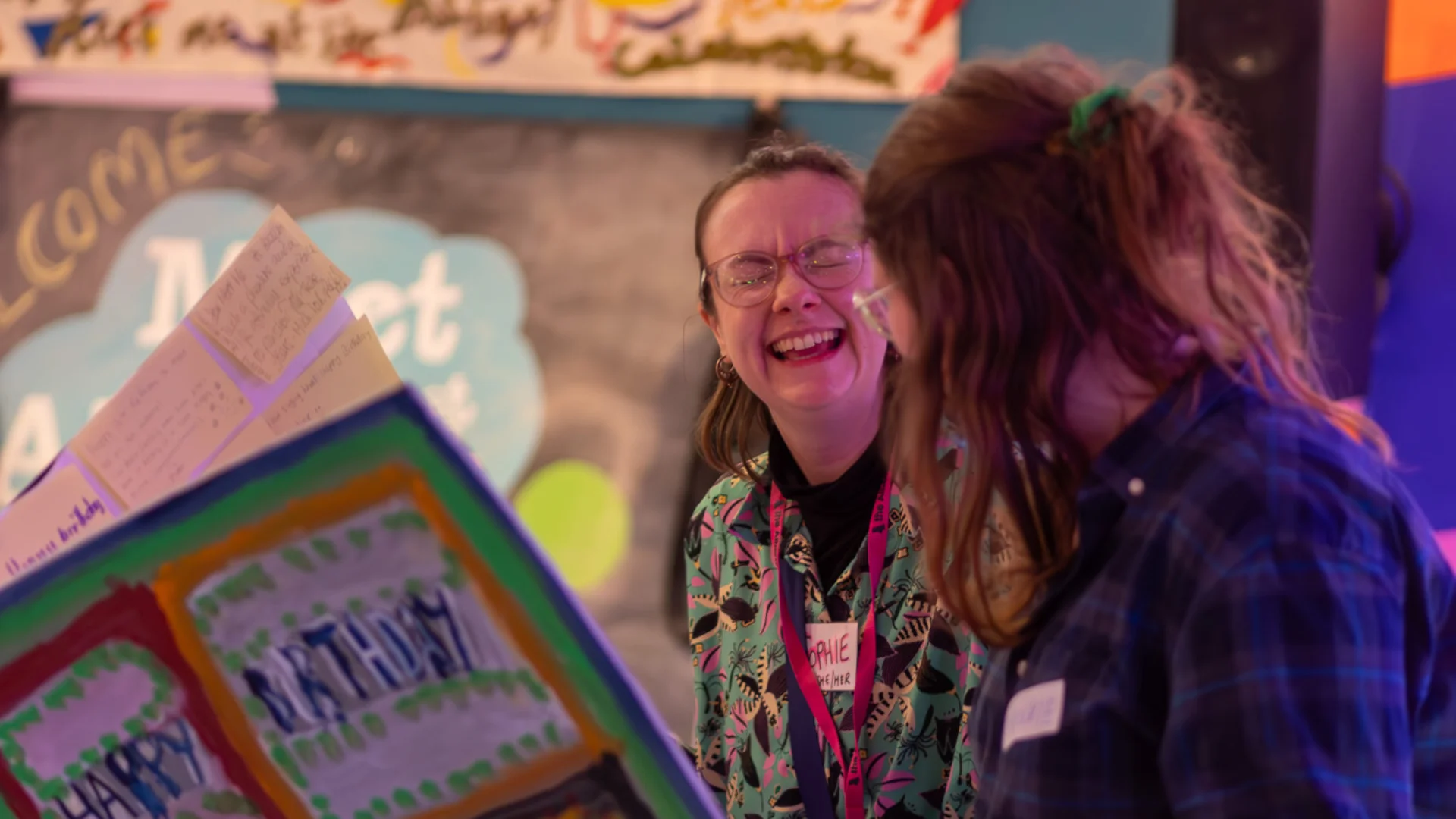 Two people laughing while looking at a comically oversized, handmade, colourful card with "Happy Birthday" on it. A festive, joyful atmosphere with a decorated backdrop.