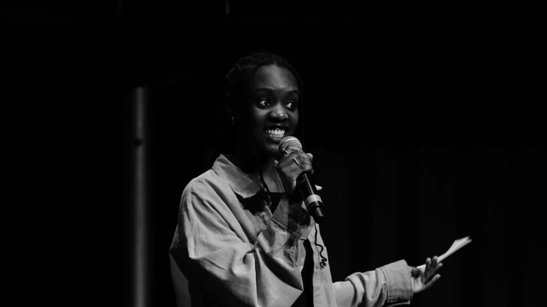A young person speaks into a microphone on stage. They wear a loose shirt, hold a small notecard, and convey enthusiasm. The image is in high contrast black and white.