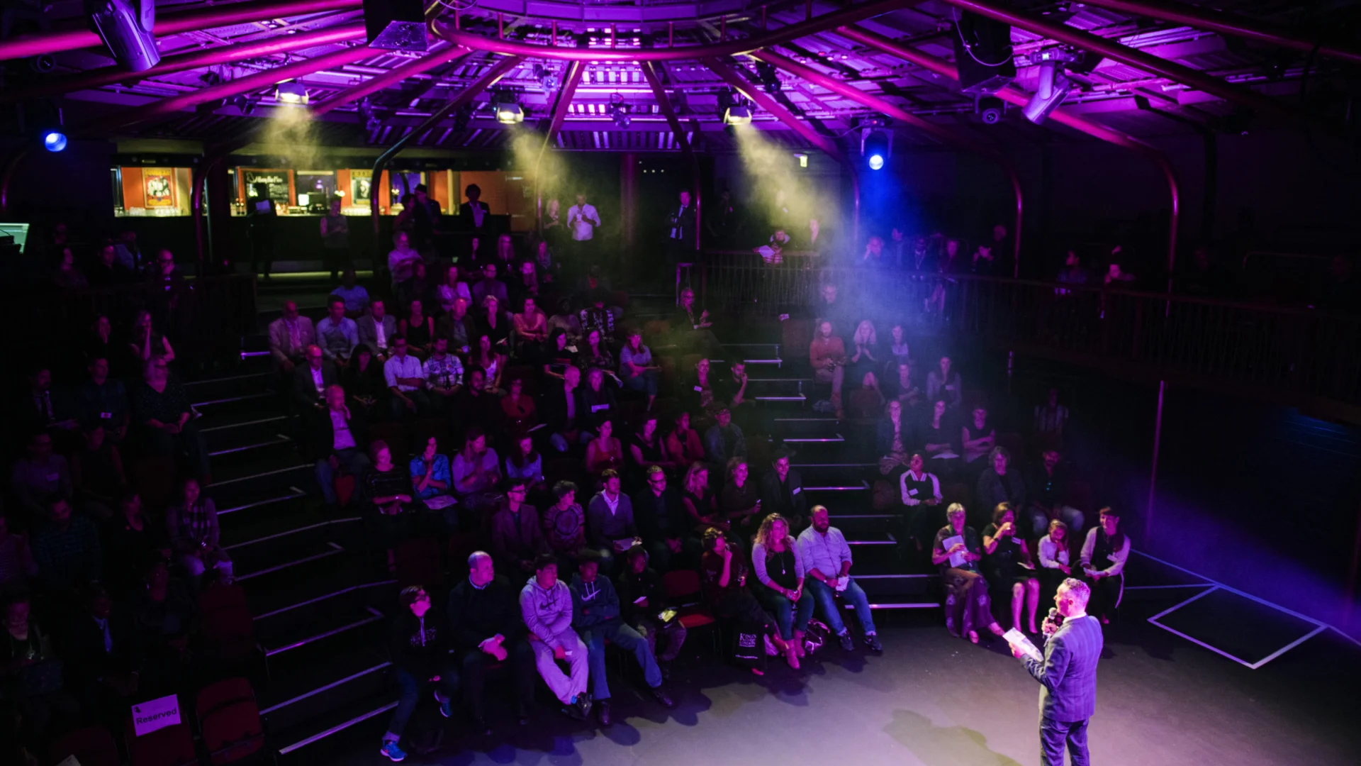 The image shows a theatre audience watching a sole presenter on stage. The spotlights are pink and vibrant. The speaker has their back to the audience and is wearing a suit, holding notes and speaking into a microphone.