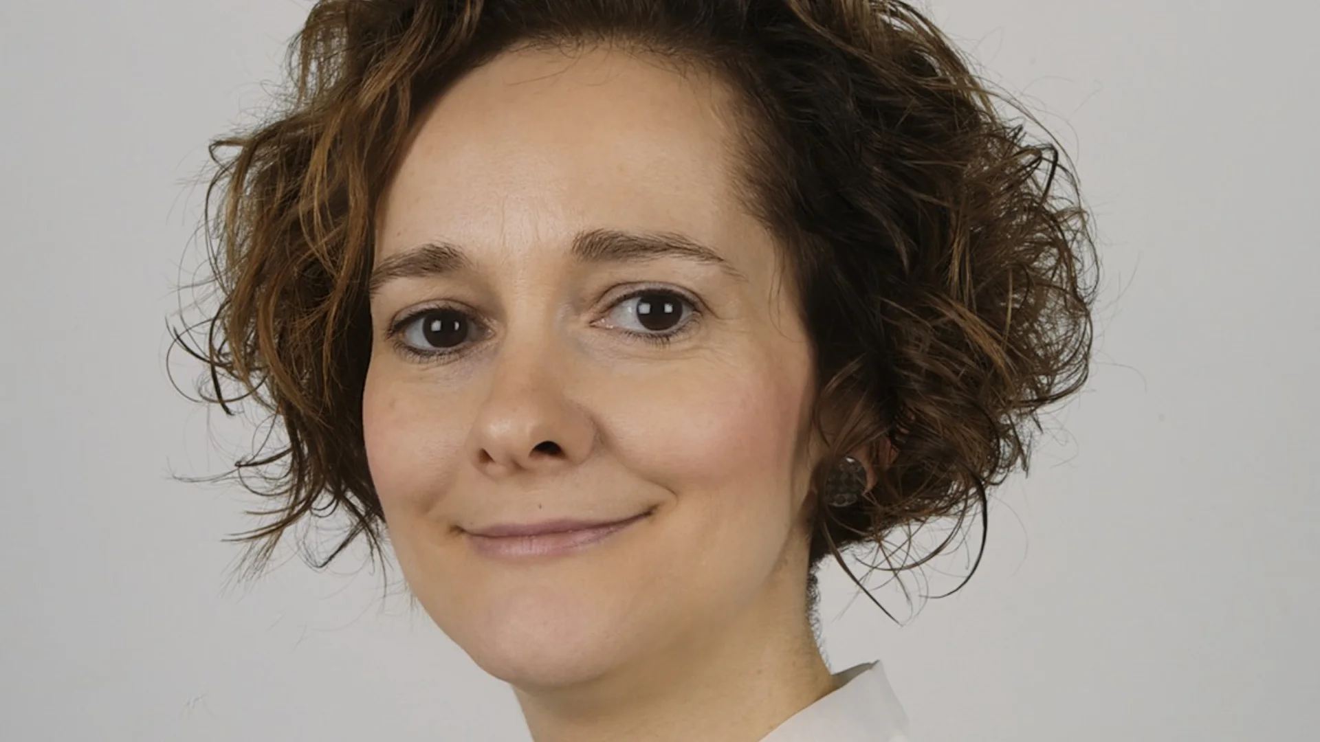 A woman with short, curly hair and a gentle smile is wearing a white blouse with a bow. The background is plain, emphasising her serene expression.