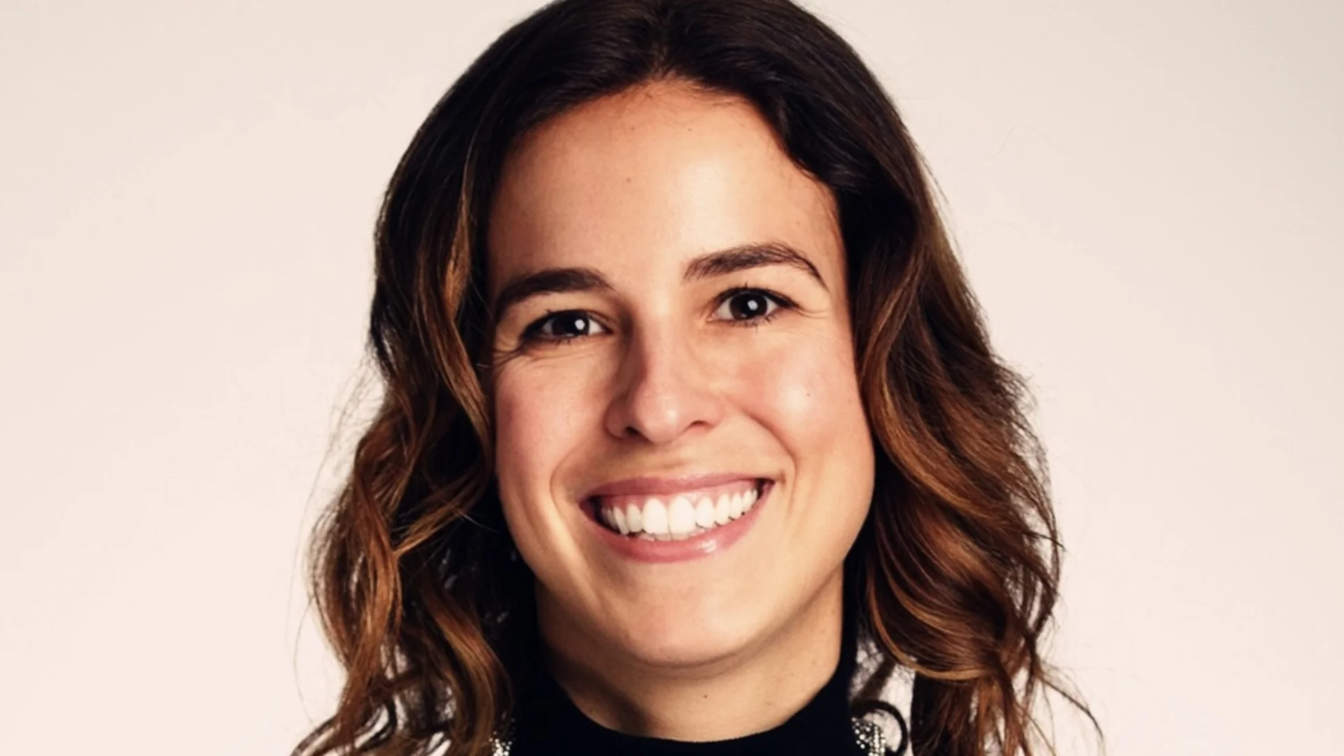 Smiling woman with wavy brown hair wearing a black turtleneck and a silver necklace. Background is plain light gray, conveying a friendly, professional tone.