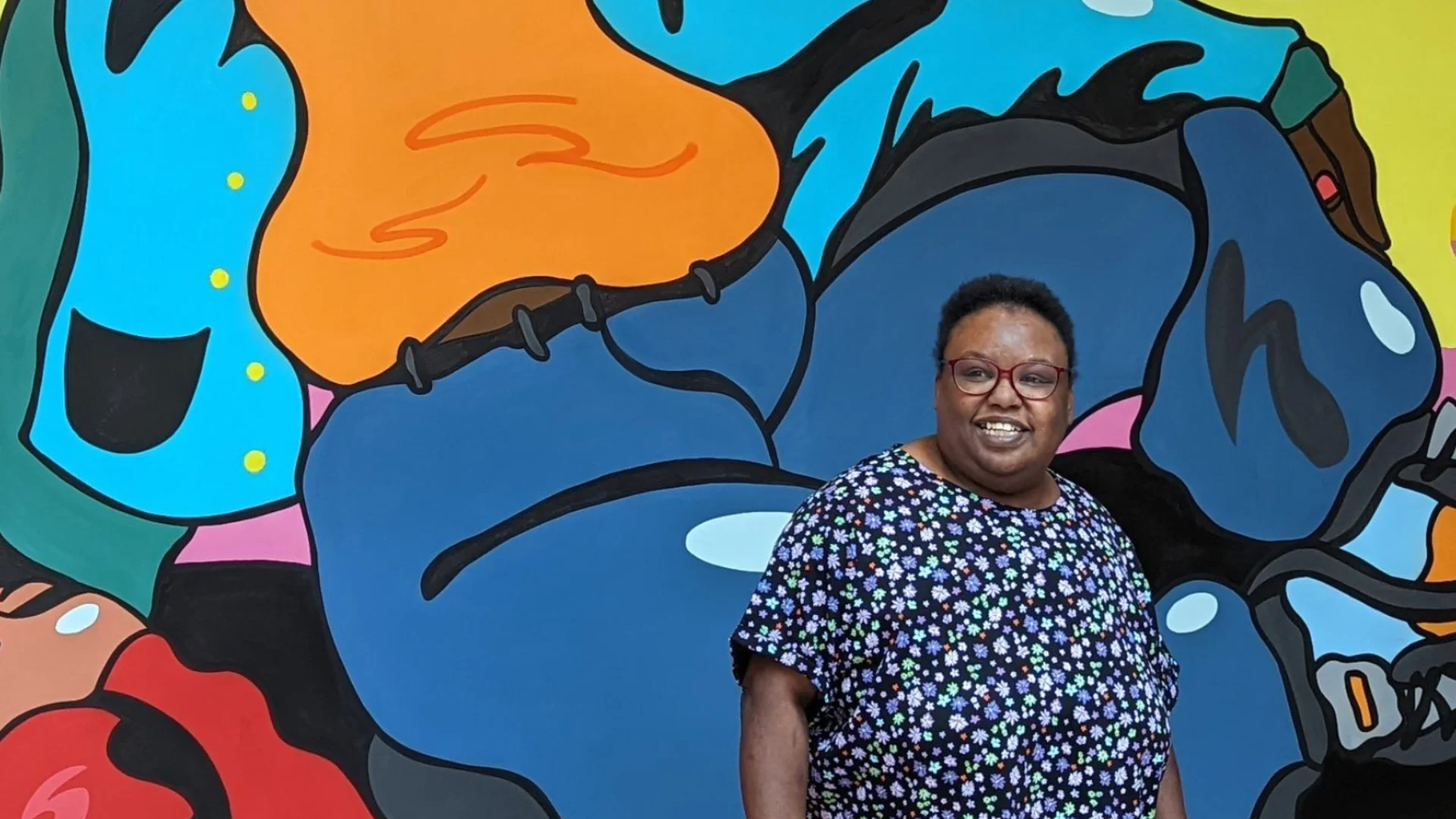 A person stands smiling in front of a vibrant mural featuring a figure with a yellow crown and a "Heart n Soul" flag, conveying joy and empowerment.