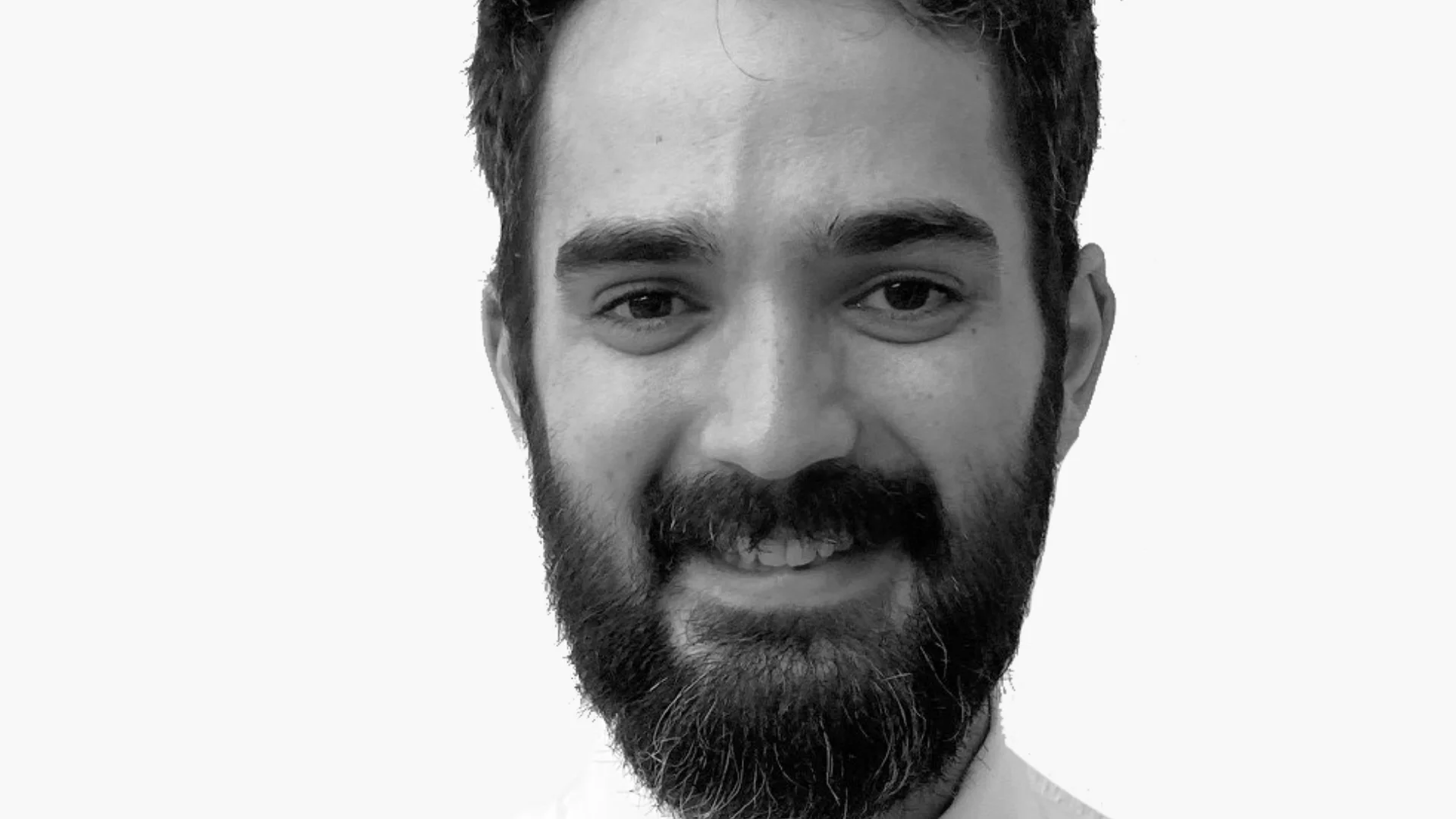 A black and white image of a smiling man with a beard in a white shirt and polka dot tie against a plain background, conveying a professional and approachable tone.
