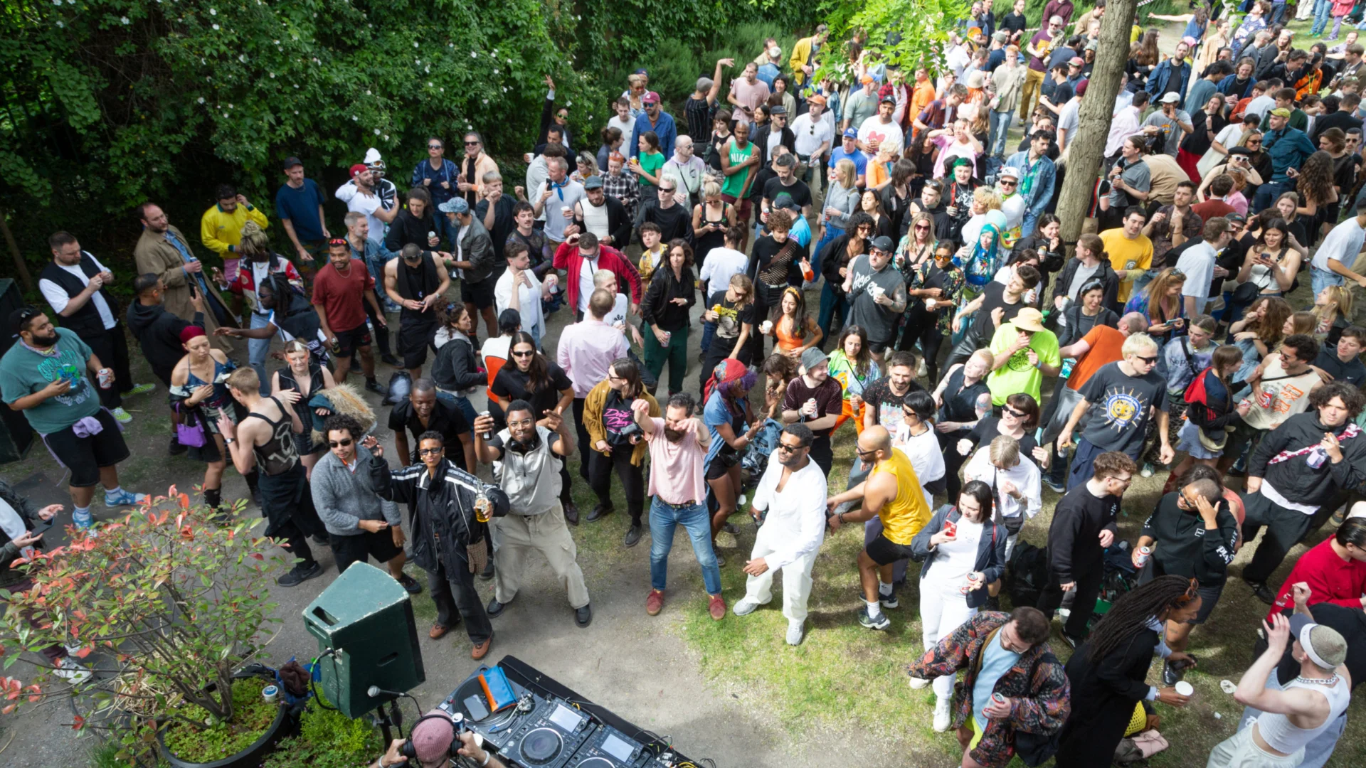 A lively outdoor party with a diverse crowd of people dancing and socialising. A DJ booth is visible, surrounded by green trees. Warm, joyful atmosphere.