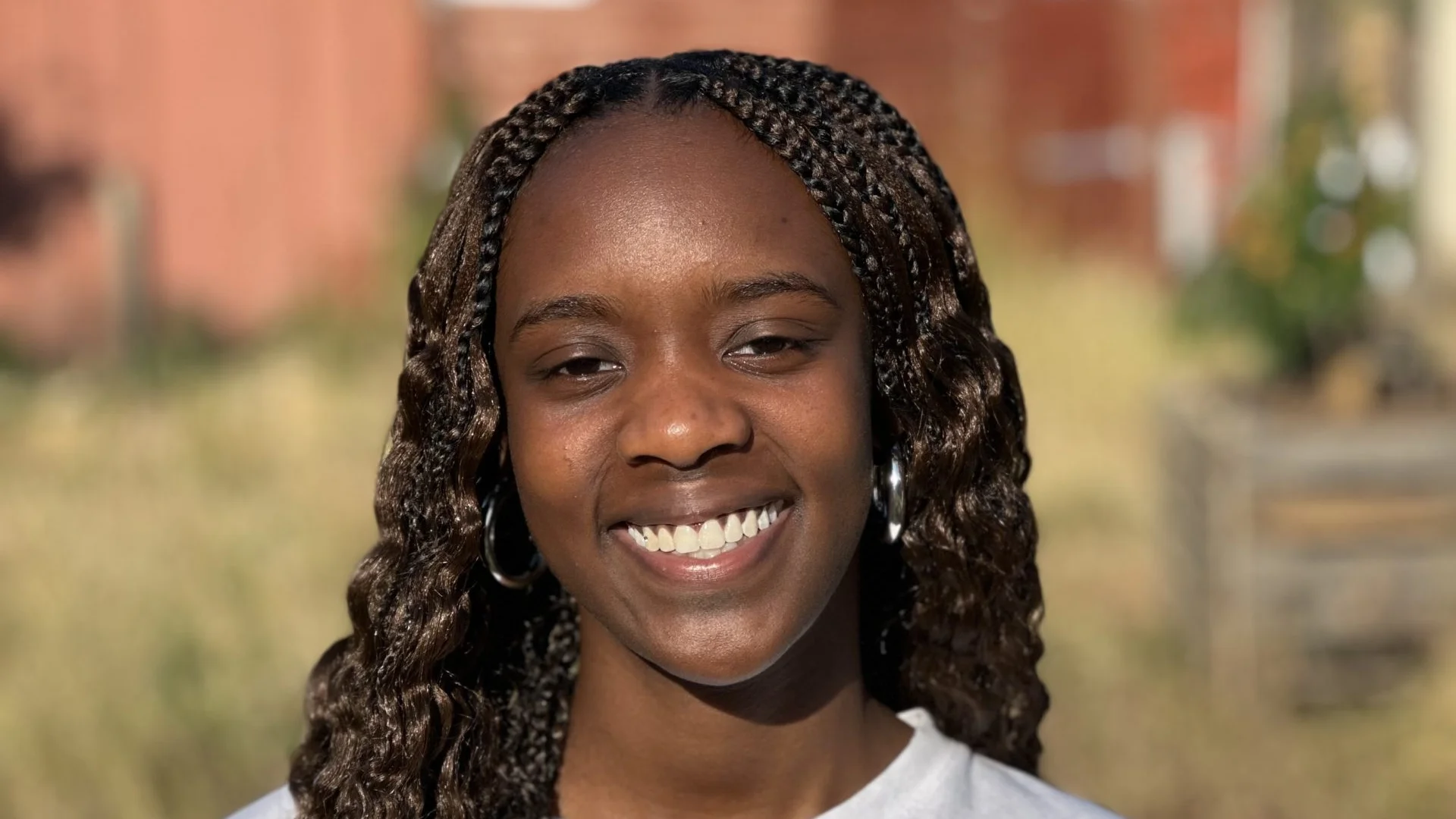 A photo of a black woman with long braids wearing a white converse t-shirt. She is stood in the Albany garden on a sunny day, smiling at the camera.