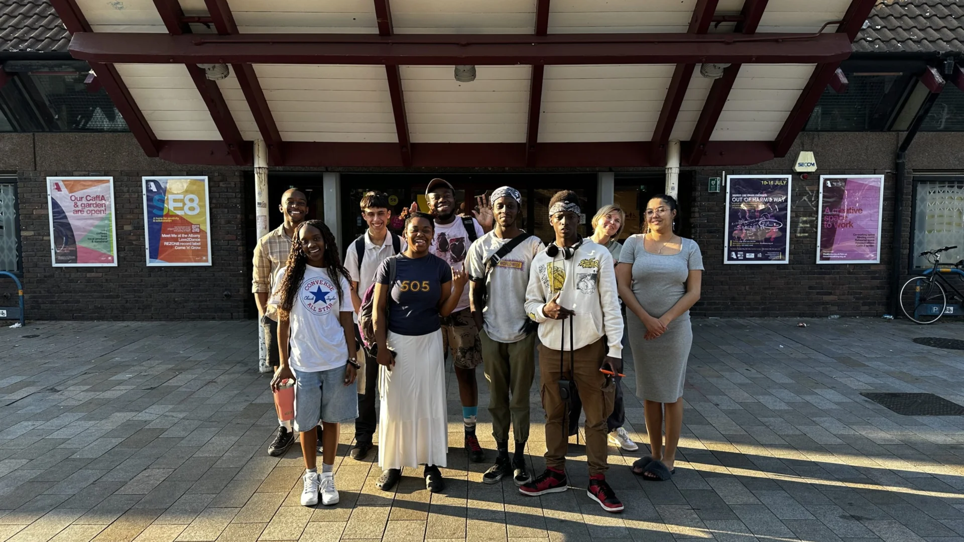 A group of young people stand smiling outside the Albany front doors. The sun is shining bright, creating a warm and friendly atmosphere.