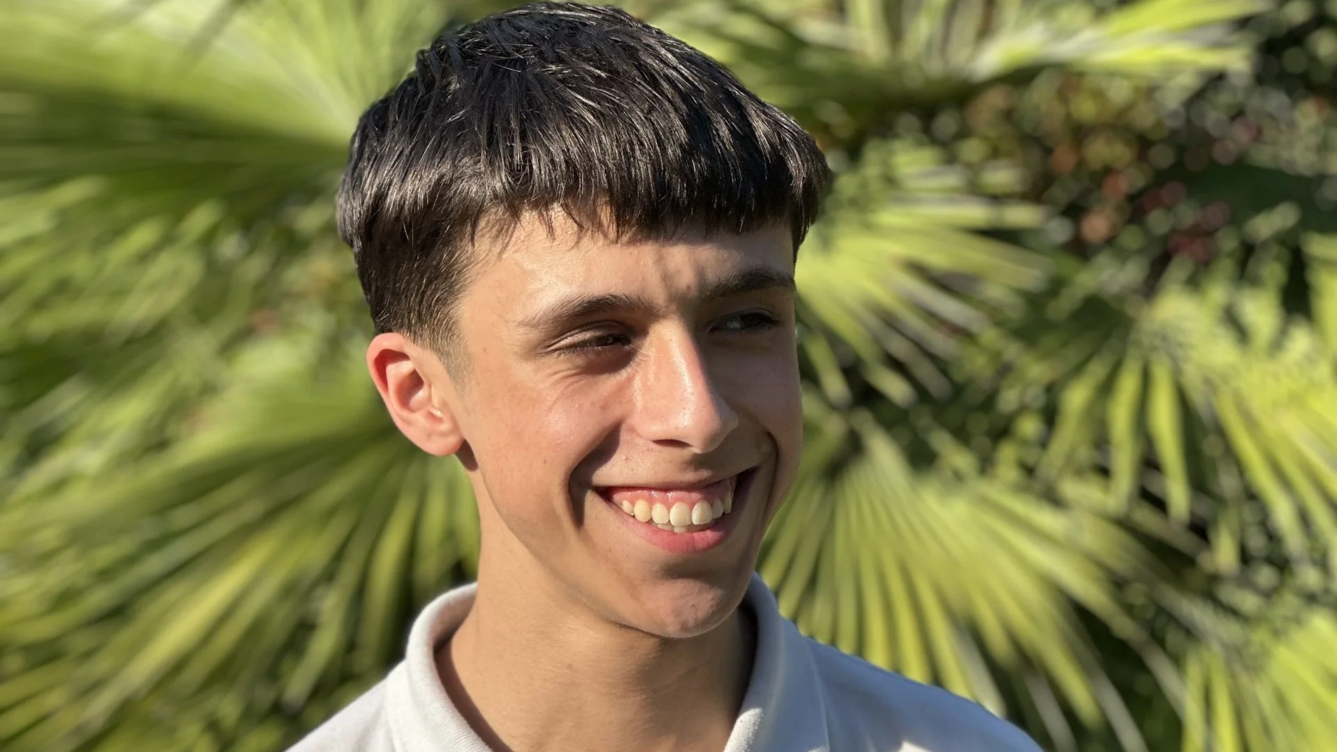 A photo of a young man with black hair and wearing a white polo shirt. He is stood in front of a palm tree on a sunny day and his eyes are squinting in the sun.