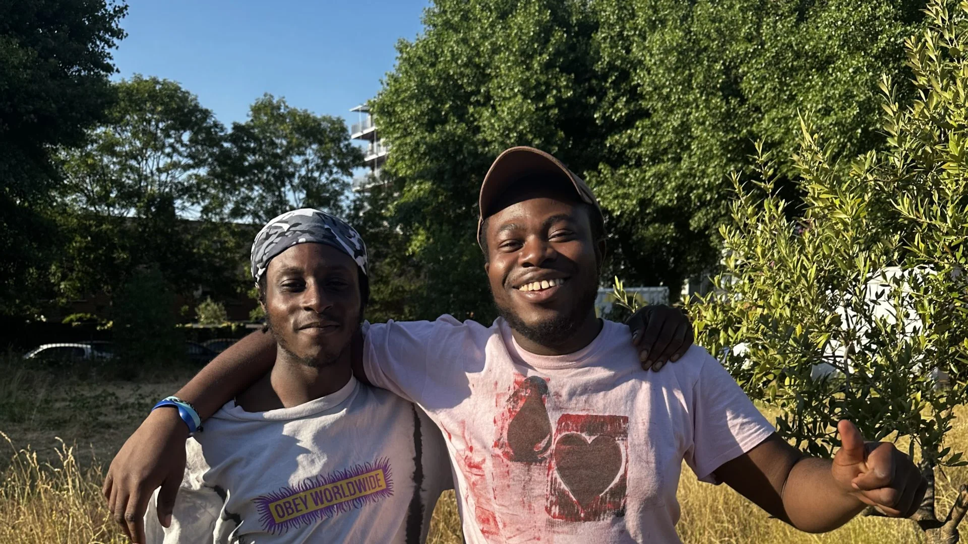 A photo of two young black men wearing t shirts and shorts, stood with their arms round each other in the Albany garden on a sunny day.