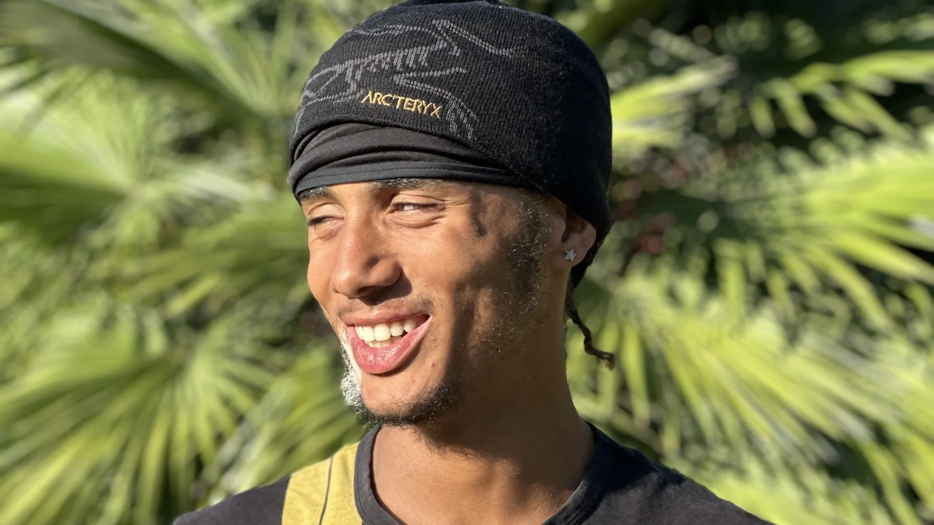 A photo of a young man wearing a black tshirt and head covering. He is stood in front of a palm tree on a sunny day and is looking to the right and smiling.