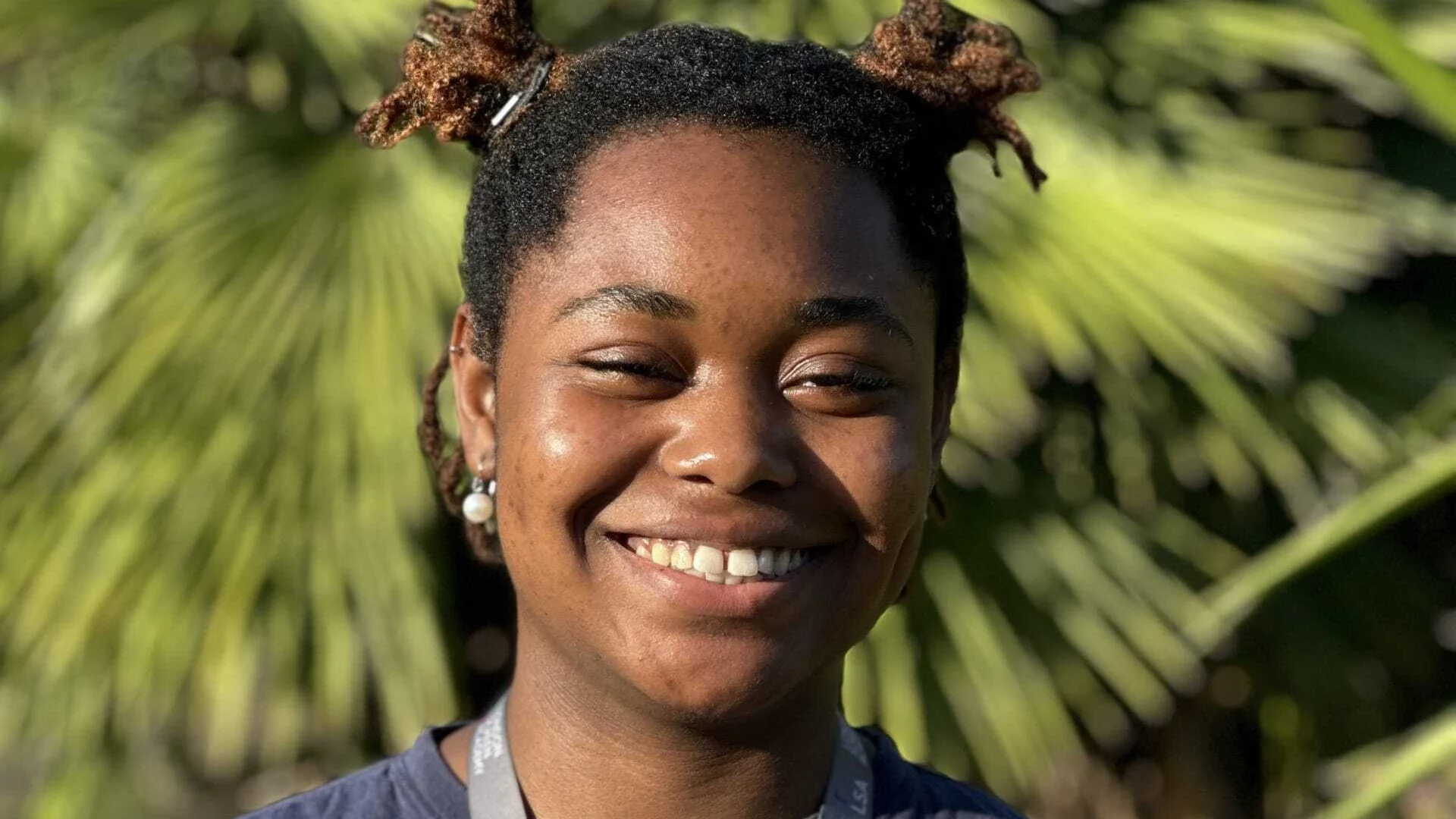 A photo of a young black girl with bunches stood in front of a palm tree on a sunny day. She is wearing a grey tshirt and lanyard and is smiling.