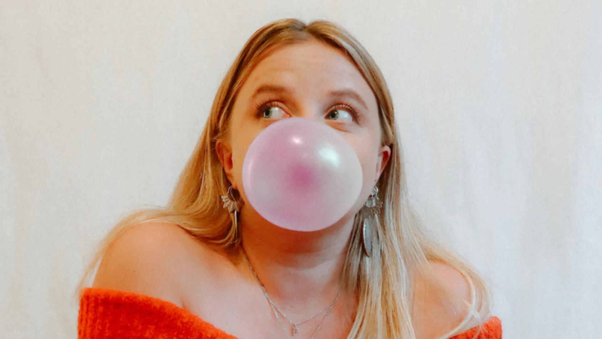 A photograph of Poppy Daniels, a blonde woman wearing an orange top and blowing a bubble of pink bubblegum.