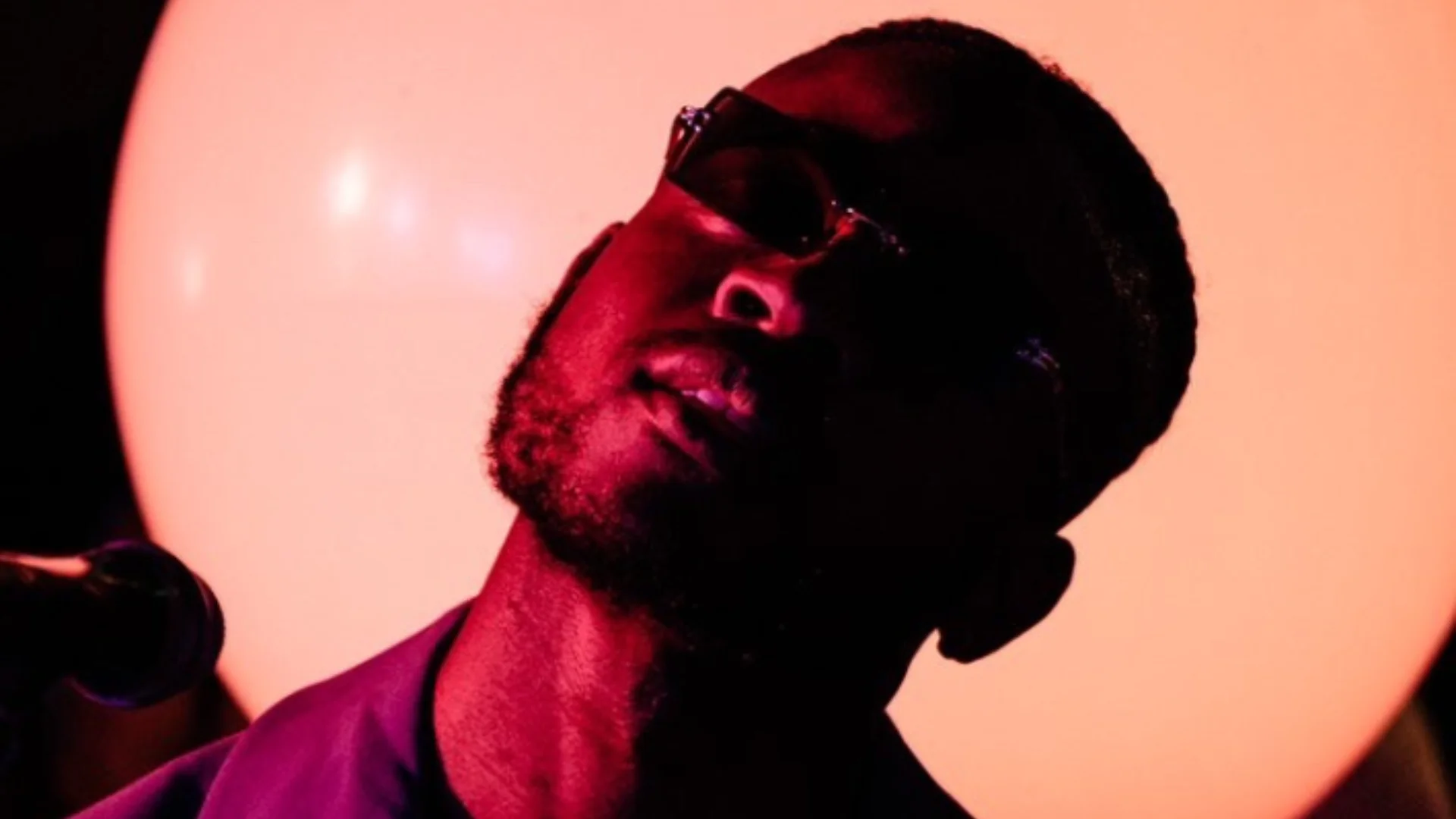 A headshot of the artist INSXGHT. A black man wearing sunglasses, in shadows, head leaning back, with an orange circle lit up behind him.