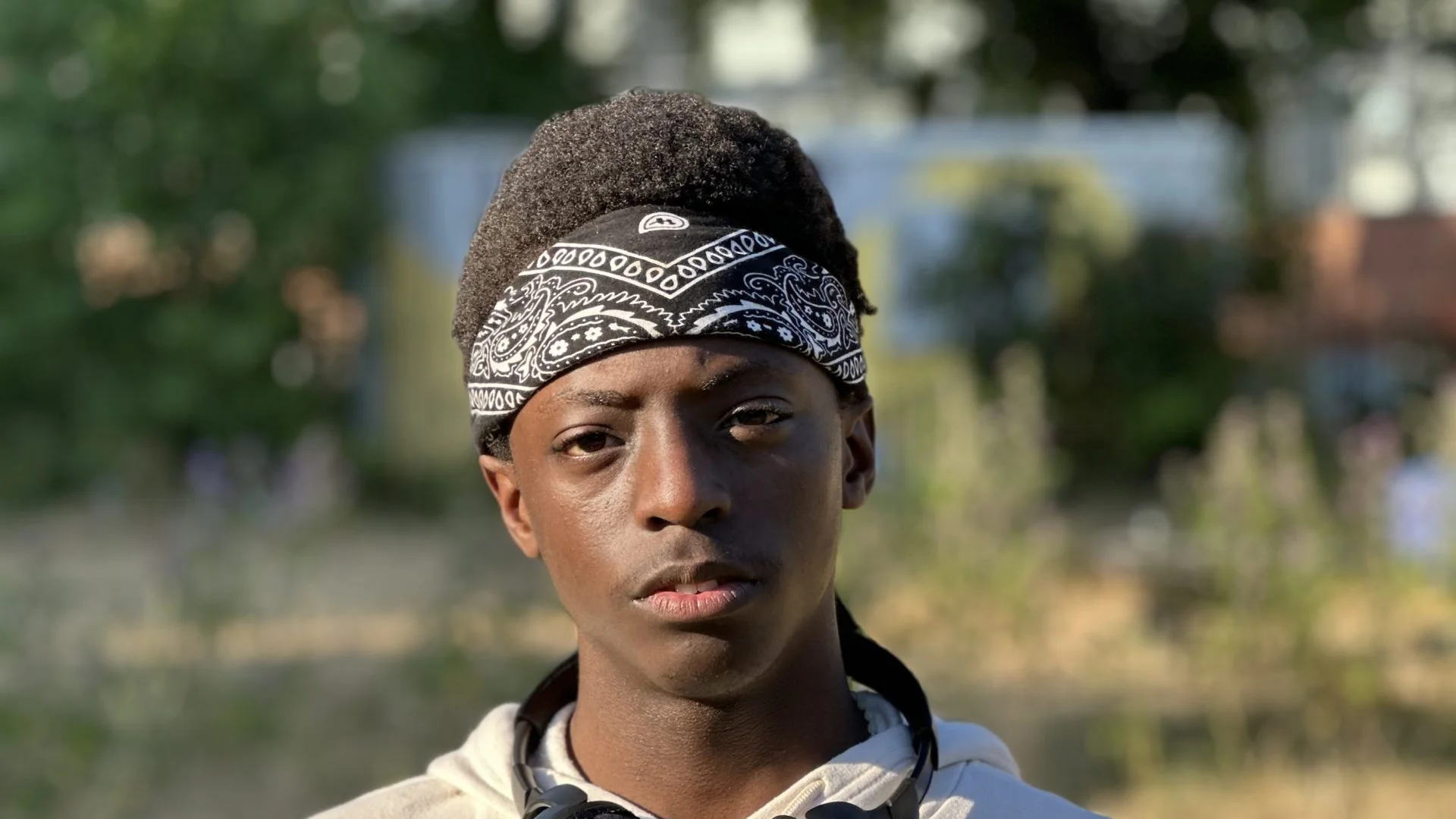 A photo of a young black man wearing a black bandanna and a hoodie, with headphones round his neck. He is stood in the Albany garden on a sunny day.