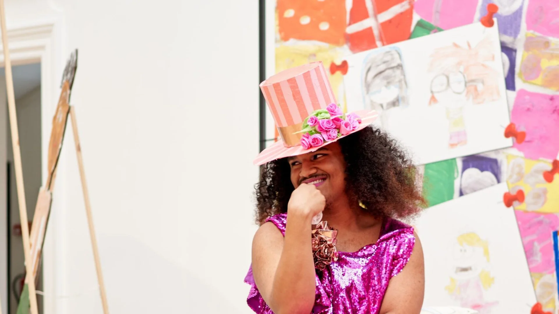 A photo of Sebastian - a person with long black curly hair, wearing a pink dress and pink top hat, sat on a bed with a contemplative expression.