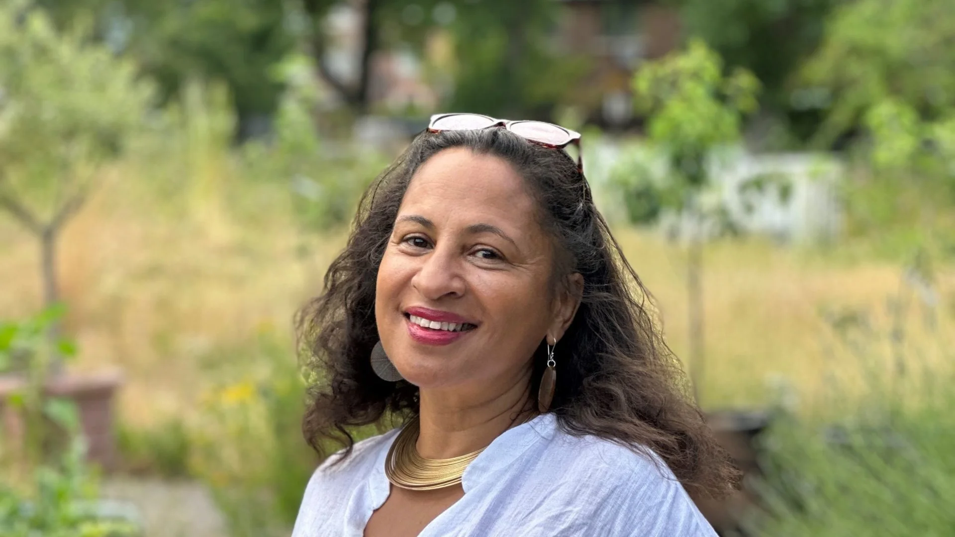 Woman in a white blouse smiles warmly in a lush garden setting. She wears sunglasses on her head and gold jewellery, creating a serene and joyful tone.