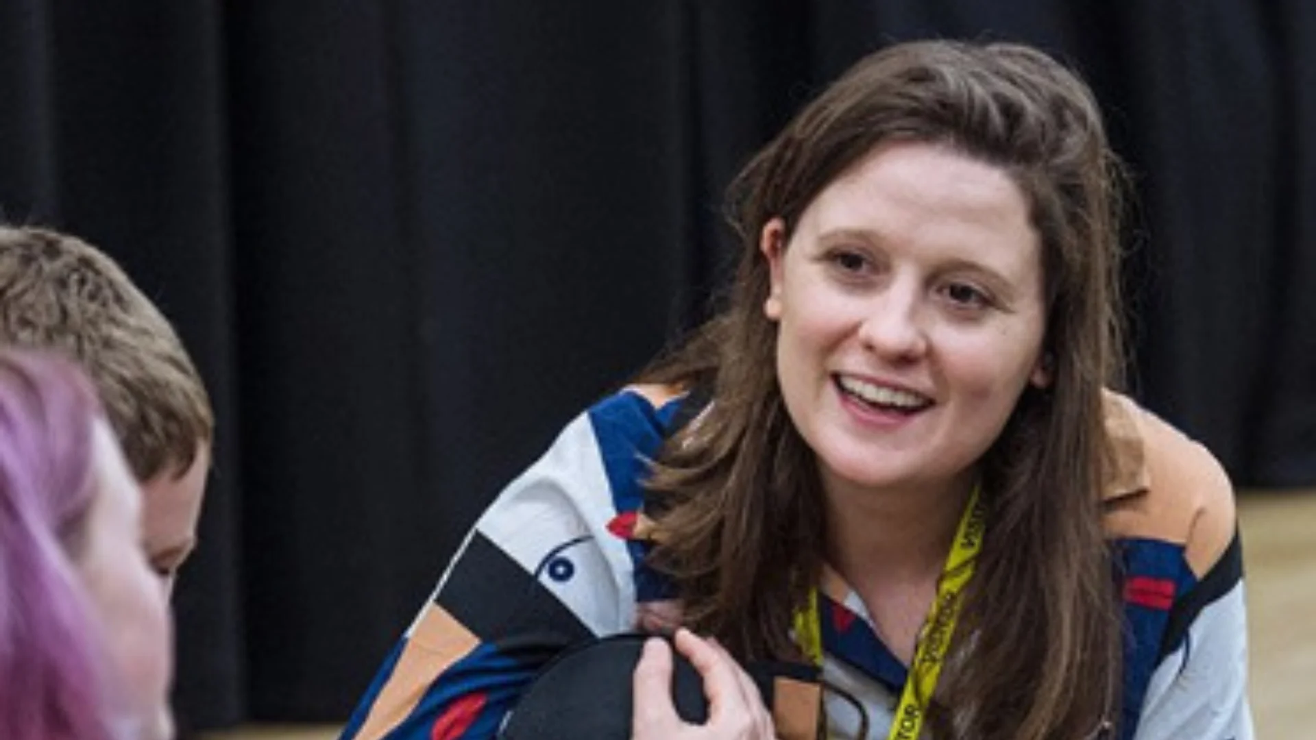 A photo of LAStheatre producer Liz Bate, a white woman with brown hair wearing a colourful shirt, kneeling on the floor.