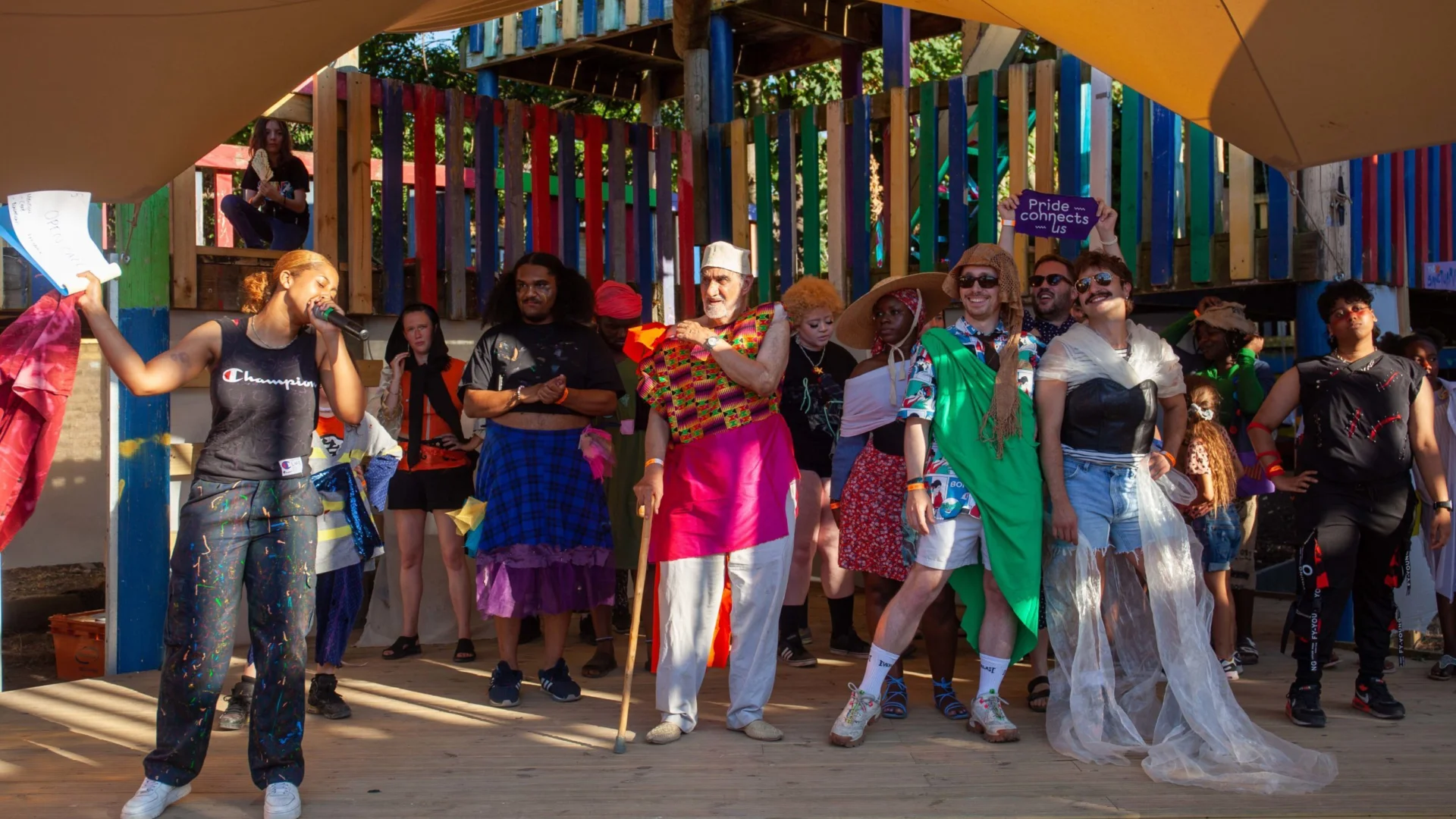 A lively group on a colourful stage celebrates, dressed in colourful, eclectic outfits. A person in holds a mic, creating an energetic, festive atmosphere.