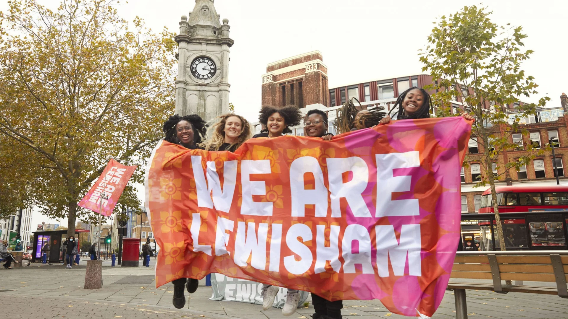 A group of smiling people jumping and holding a large banner reading "We Are Lewisham" in front of a clock tower. The scene is lively, with trees and buildings nearby.