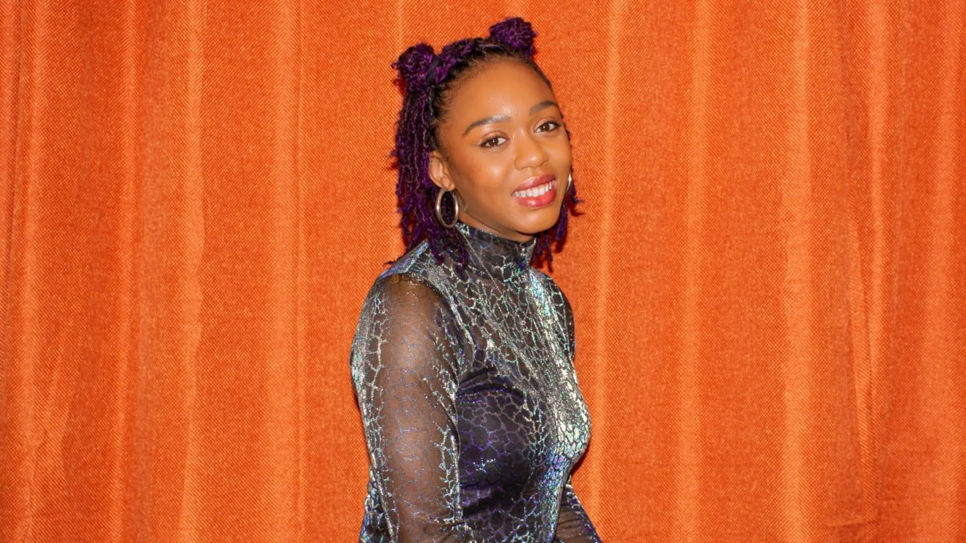 A smiling person with purple hair in a stylish updo of two small buns on top of their head, wears a shimmering, patterned dress. They stand against an orange curtain backdrop, exuding confidence.