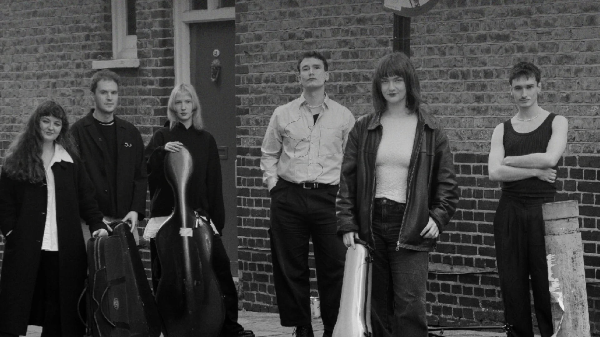 A black and white photo of a group of musicians stood on the street in front of a brick wall.