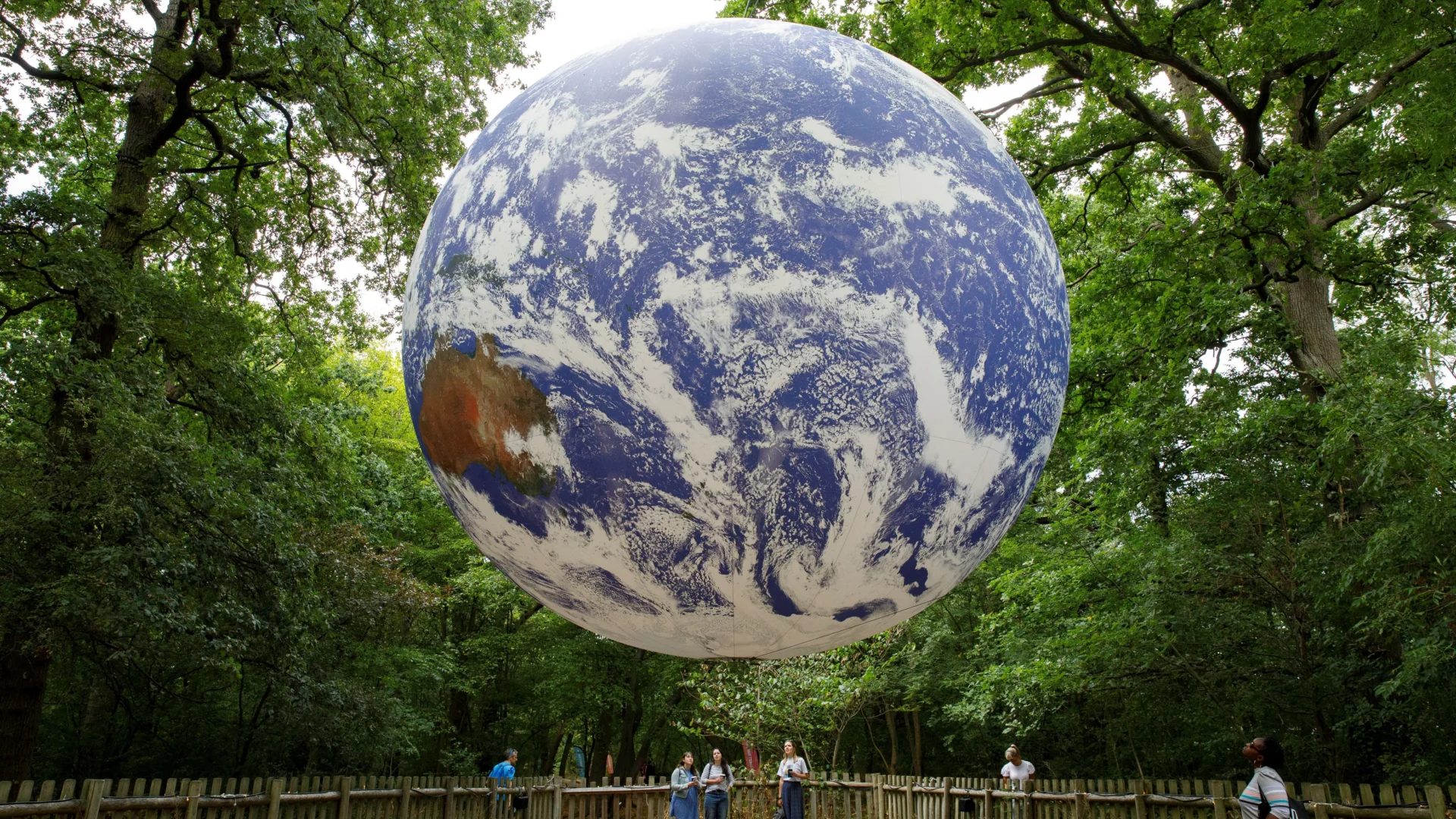 A photograph of a giant installation of the earth, created by artist Luke Jerram, suspended from some trees in a park. Underneath are some people looking up at it.