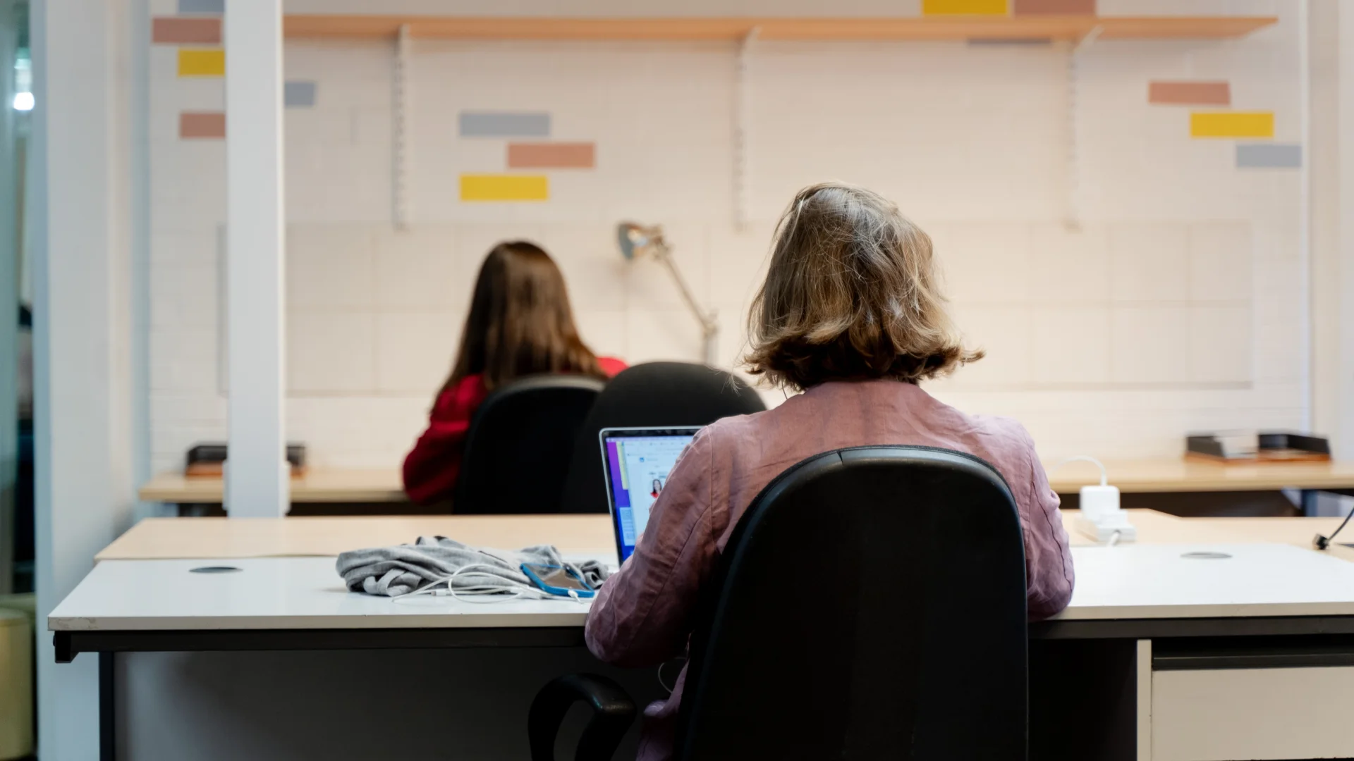 A photo of a woman with a bob working at a desk. Her back is facing the camera and she is working on a laptop with a pile of cables next to her.