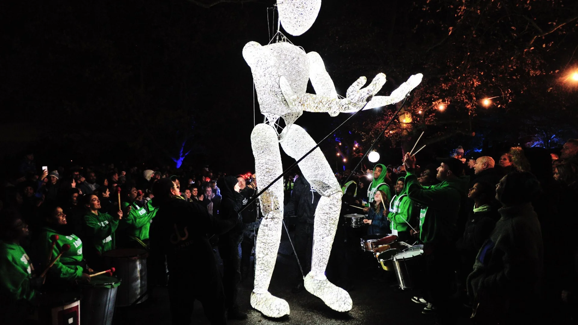 A photo of a giant human puppet lit up in bright white light. It is nighttime and through the dark you can just glimpse people in green t-shirts surrounding the puppet and drumming, and crowds of onlookers behind them.