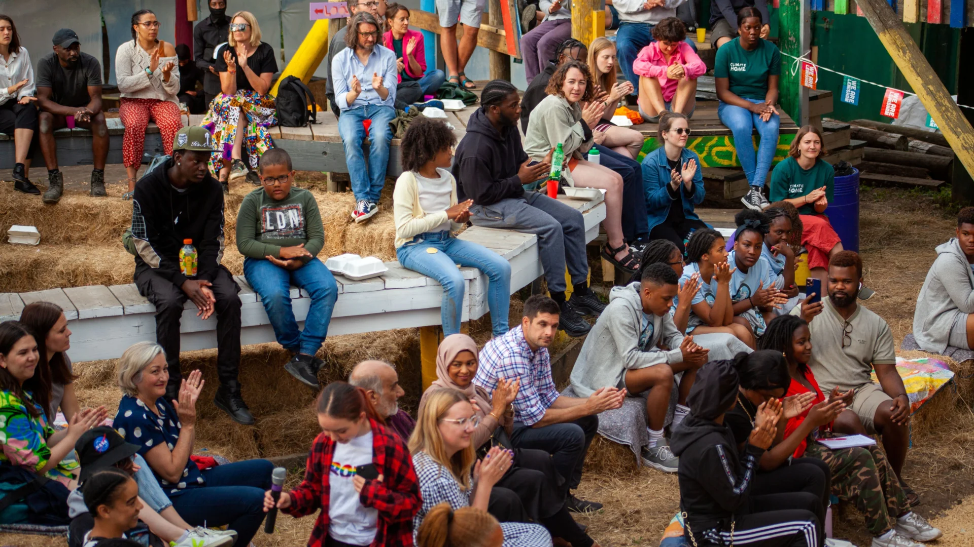A photo of a large group of people sat on hay bales in the middle of a colourful playground. They are all smiling and clapping.