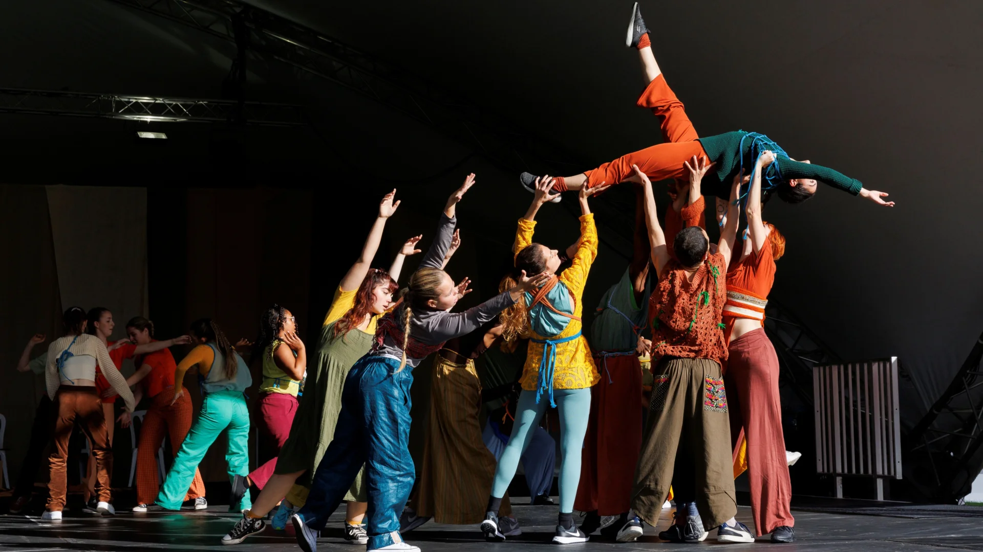 A group of dancers wearing brightly coloured clothes dance on stage. They are stood in a group with their arms outstretched, all holding up one dancer who is lying on her back with one leg pointed in the air.
