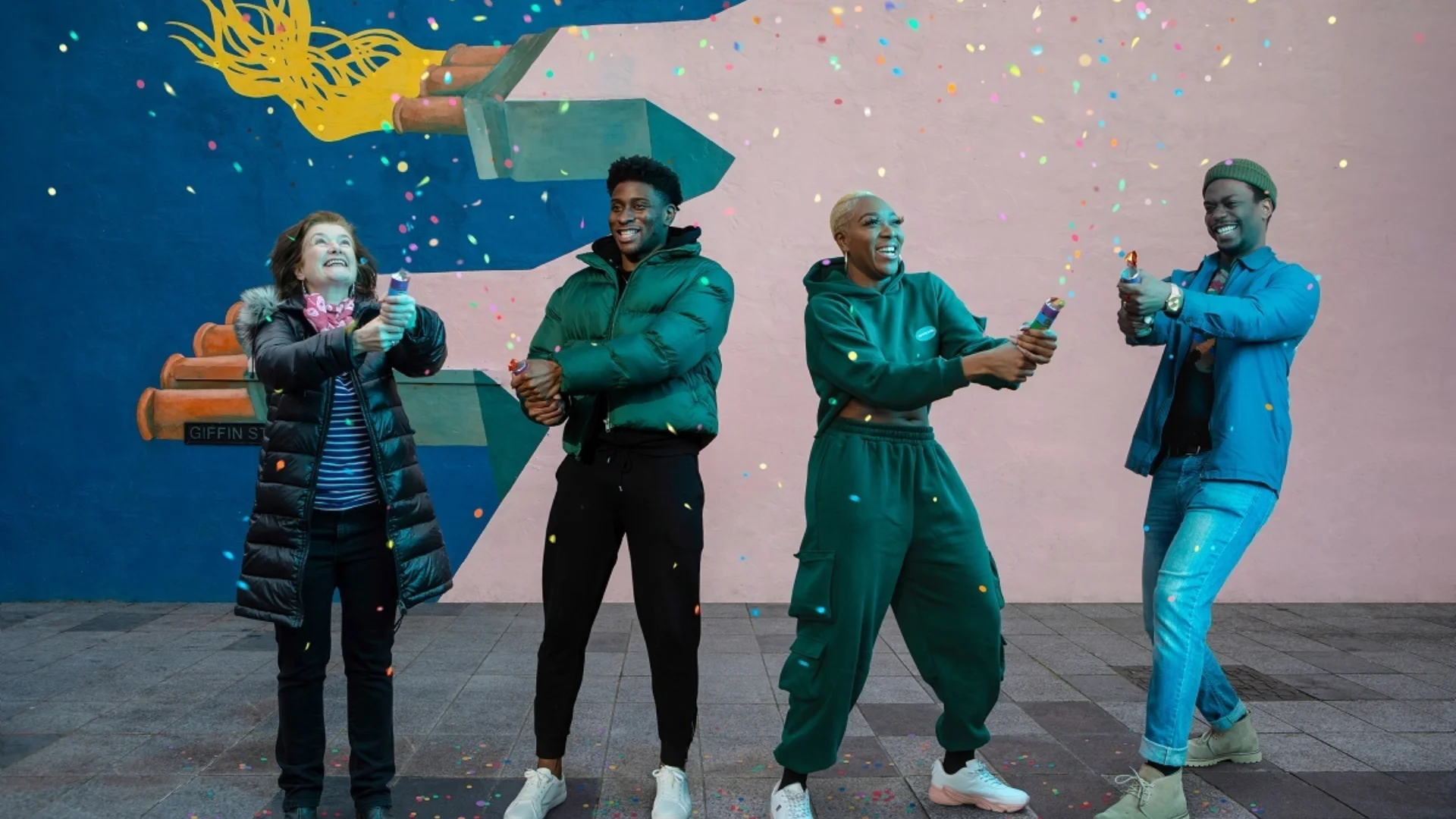 A photo of four people (Angela Clerkin, Wofai, DK, Ryan Calais Cameon) stood in front of a colourful mural in Deptford, holding confetti canons.
