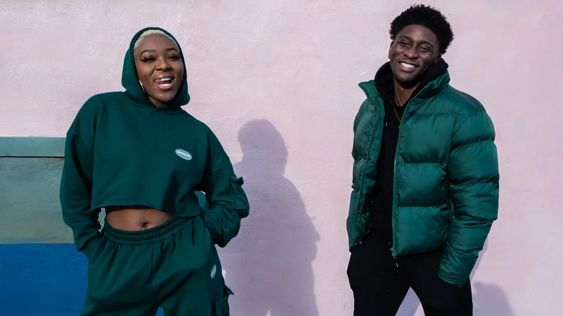 A black woman with short blond hair wearing a green hoodie with the hood up, and a black man wearing a green puffer jacket with his hands in his pockets, grin at the camera.
