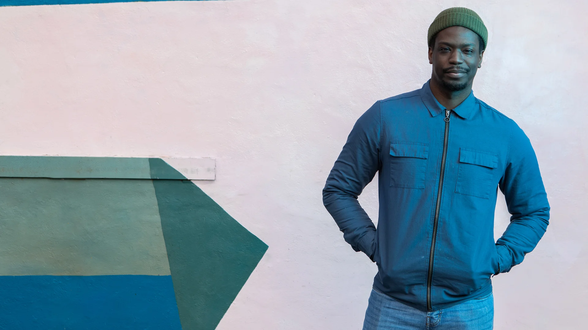 Ryan Calais Cameron - a black man wearing a green beanie hat, blue jacket and blue jeans, stands with his hands in his pockets, staring towards the camera.