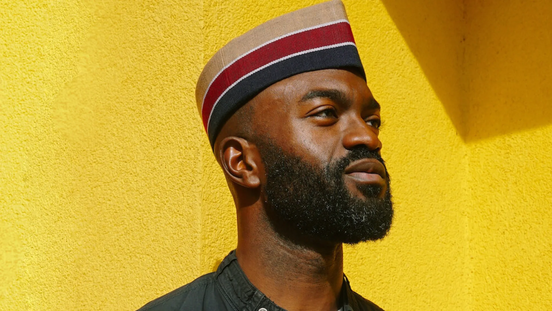 Inua Ellams - a black man wearing a red and black hat and a black jacket, stands in front of a bright yellow wall. He is gazing off to the right of the camera.