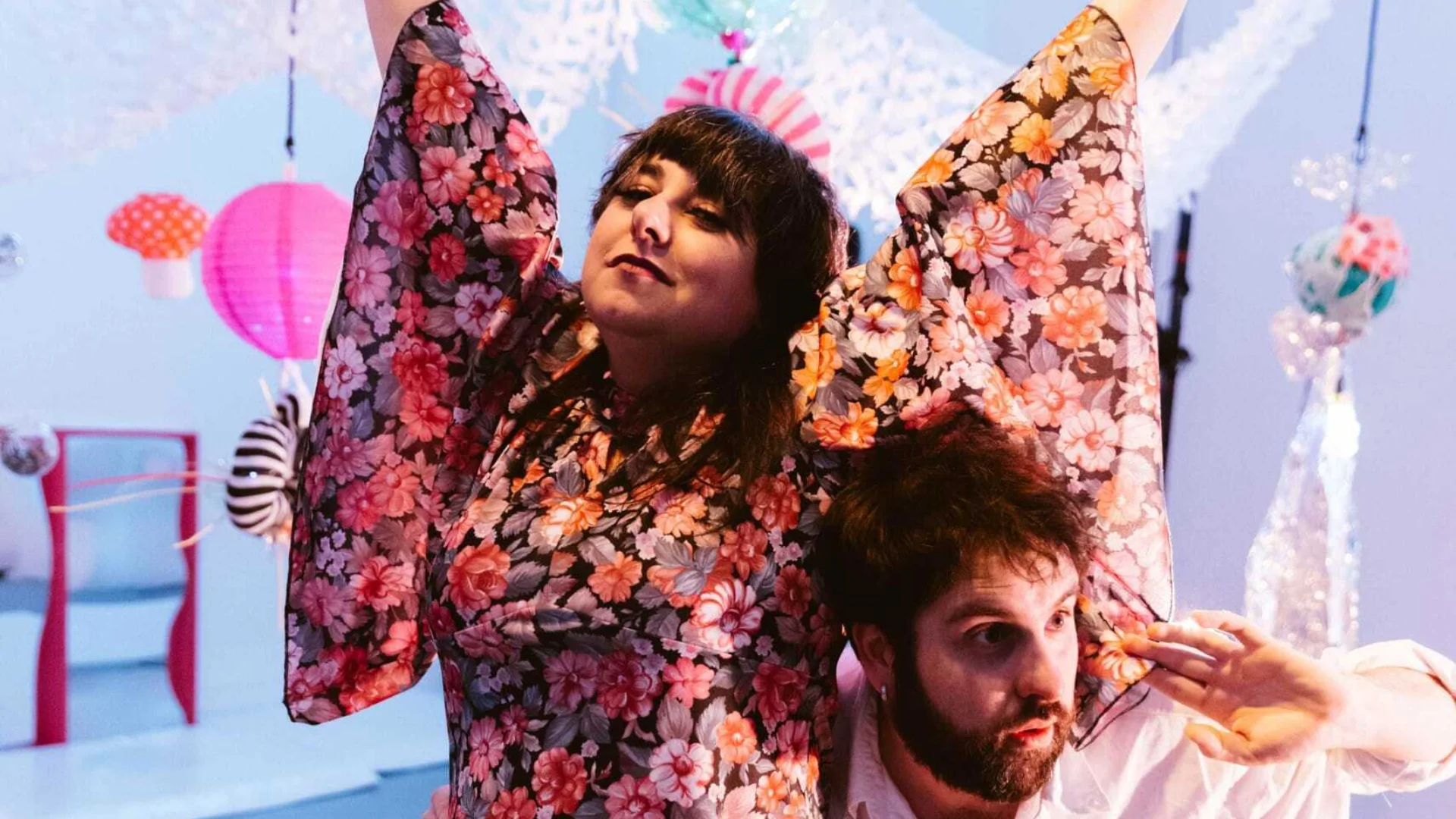 A woman with long brown hair wearing a red floaty dressed has her arms raised in the air. Peeking out behind her is a man wearing a white shirt. Behind them are colourful lights and cushions.