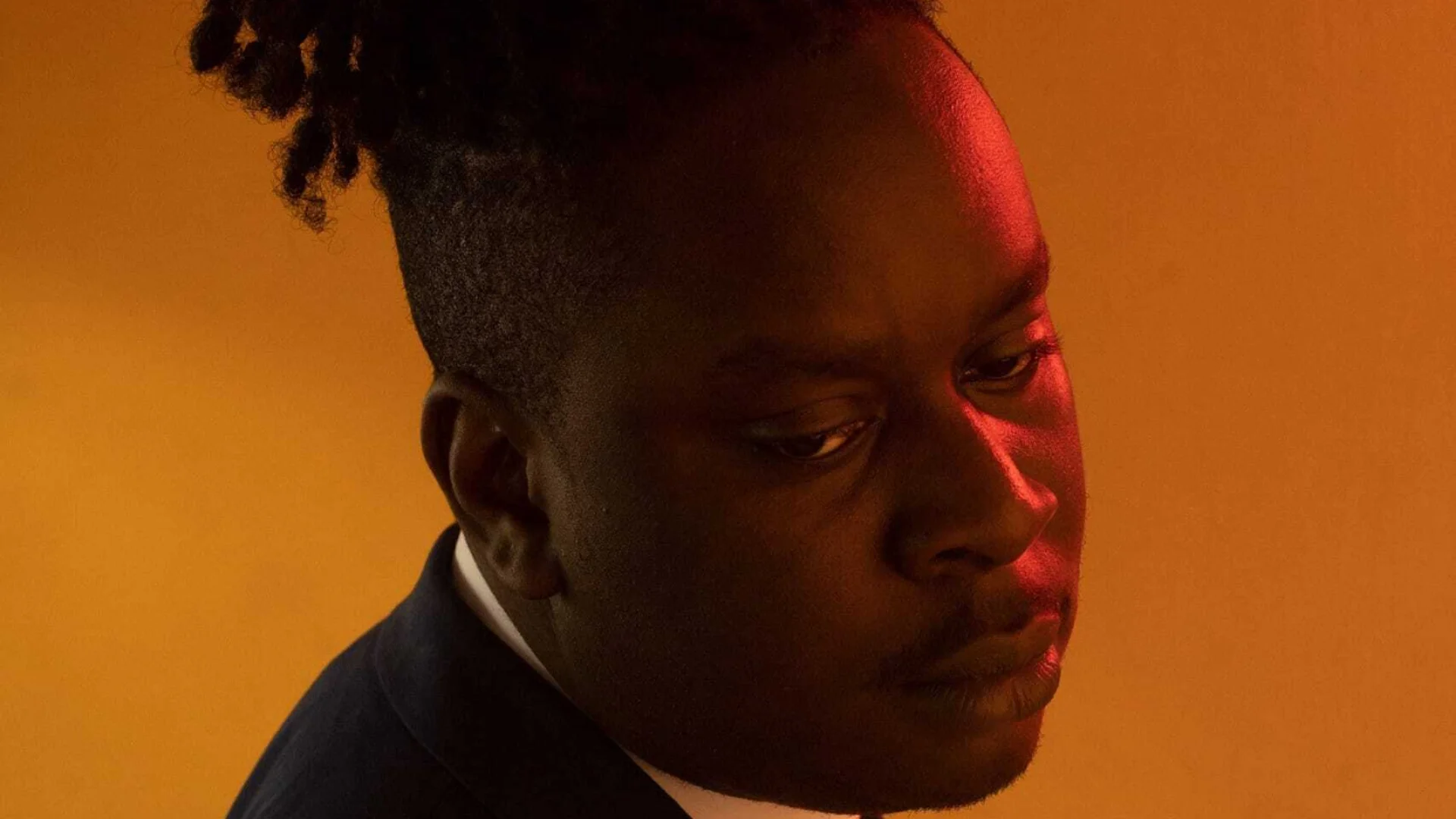 A photo of a black man with braids piled on his head. He is wearing a suit and gazing contemplatively down. The photo is artistic and bathed in orange light.