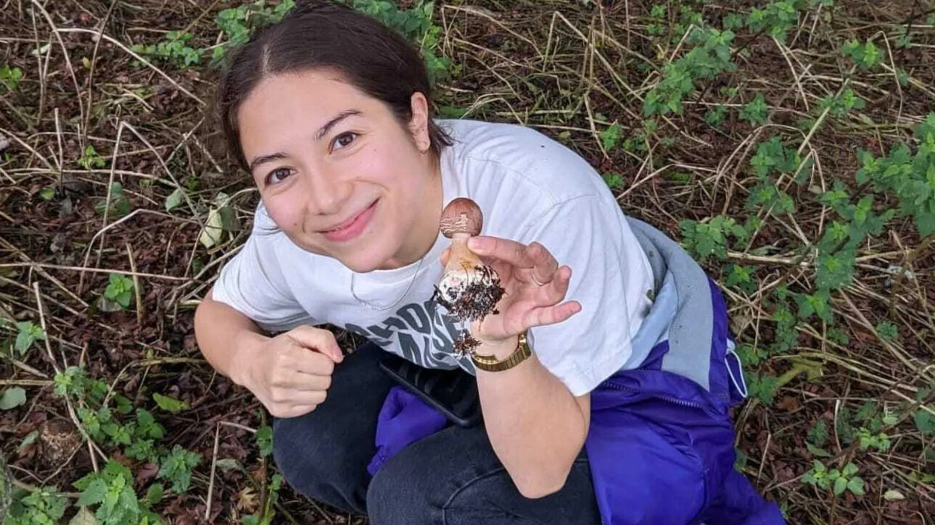 A photo of a person wearing blue trousers and a white t-shirt, crouched in a forest holding a mushroom up to the camera.