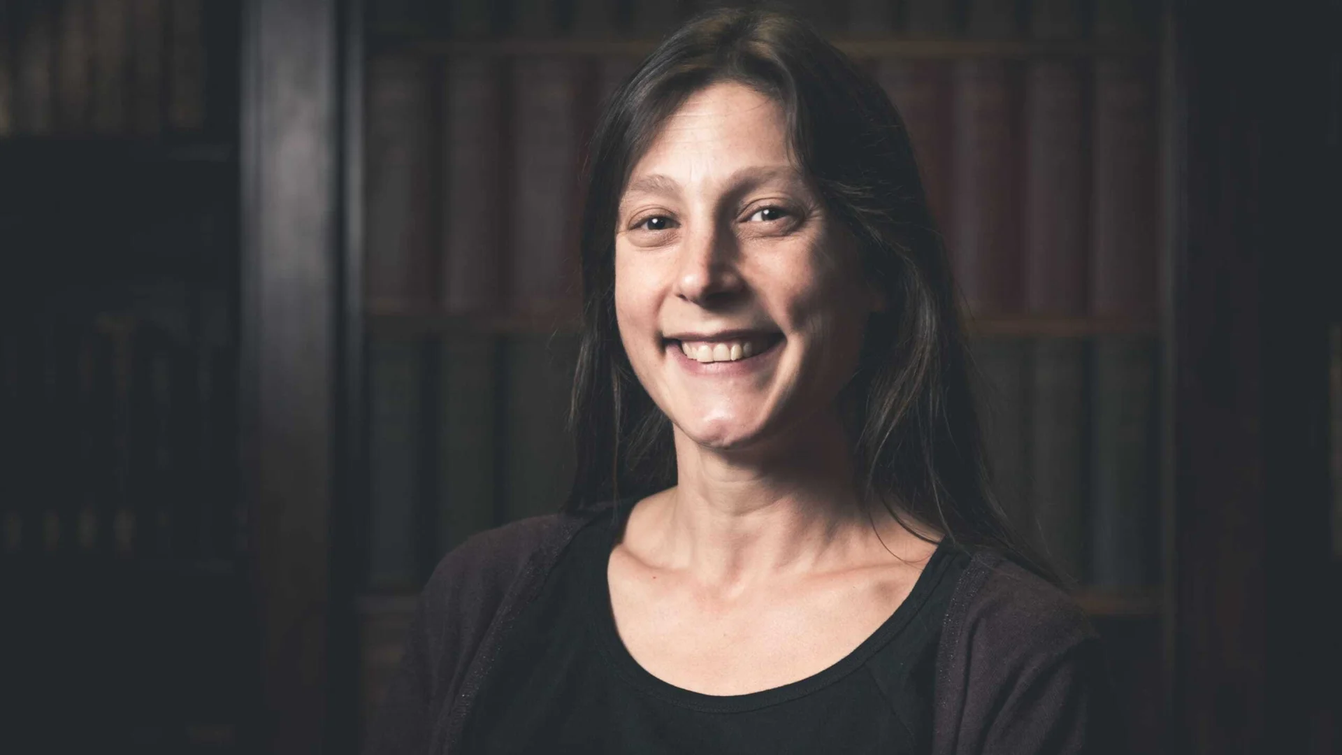 A photo of a woman with long dark hair, wearing brown hair and stood against a brown background. She is smiling at the camera.