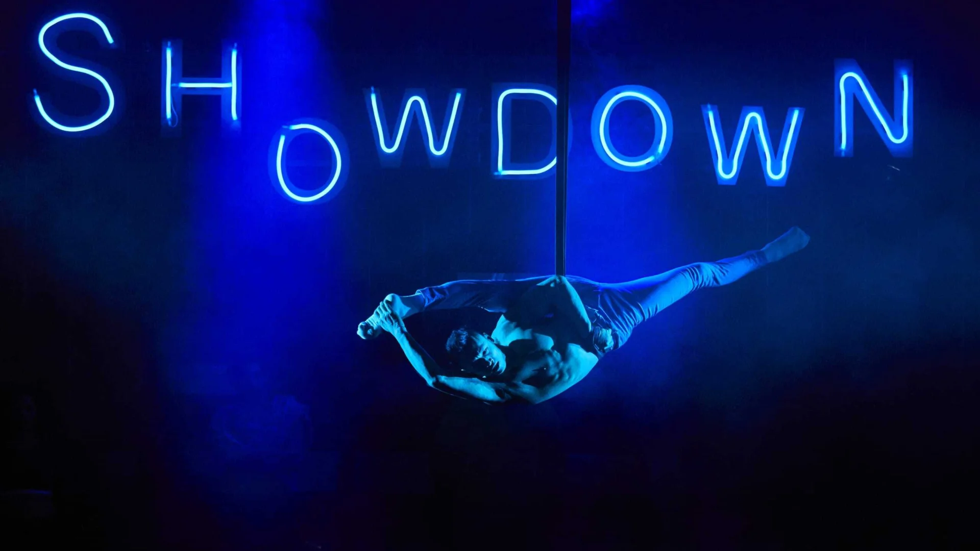 A photo of a performer suspended from a wire in the middle of a stage. Behind them the word 'Showdown' is illuminated in blue LED lighting.