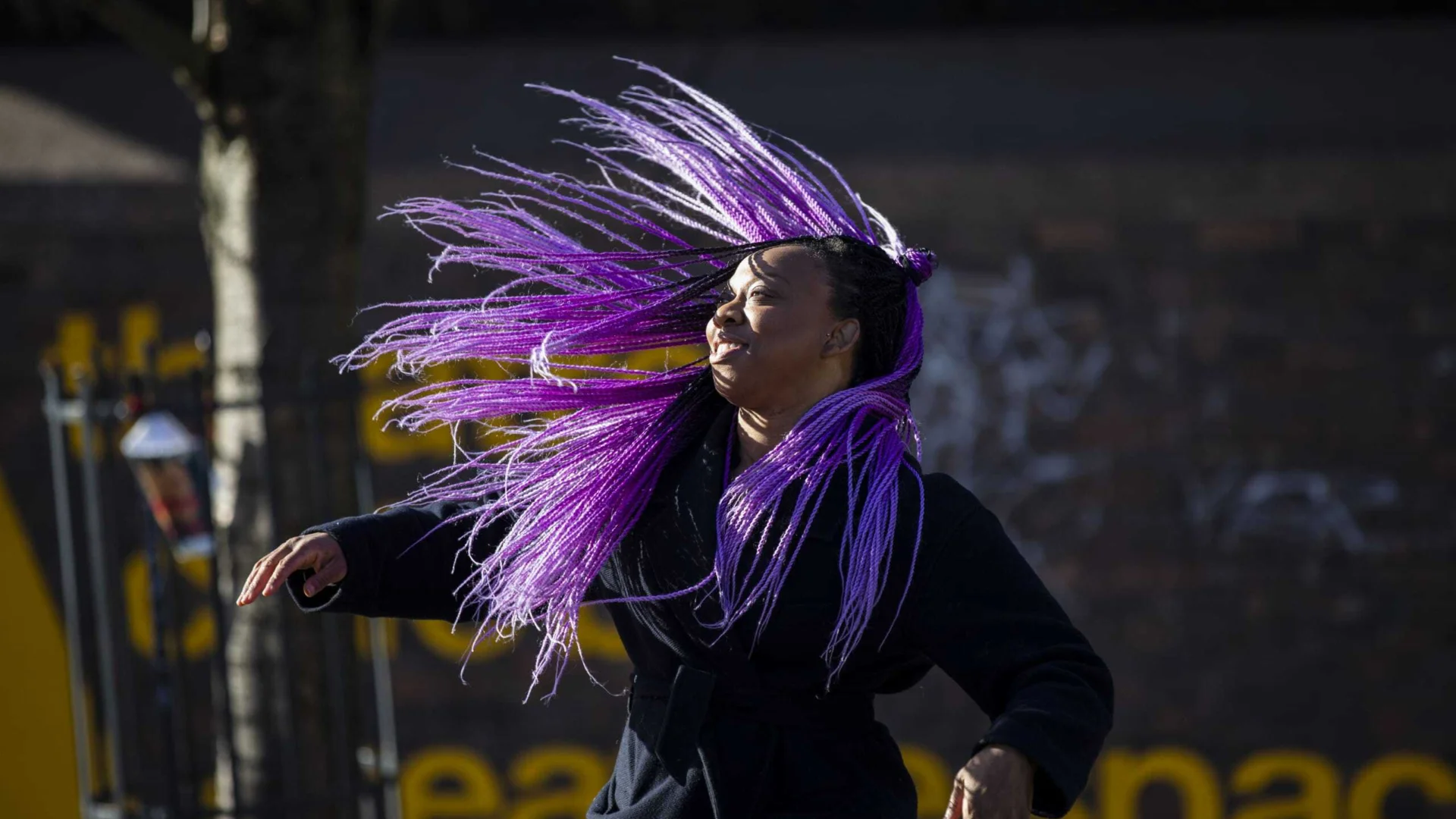 A photo of Vicki Dela Amedume, a black woman with long purple braids. She is stood outside the Albany and whipping her braids around her.